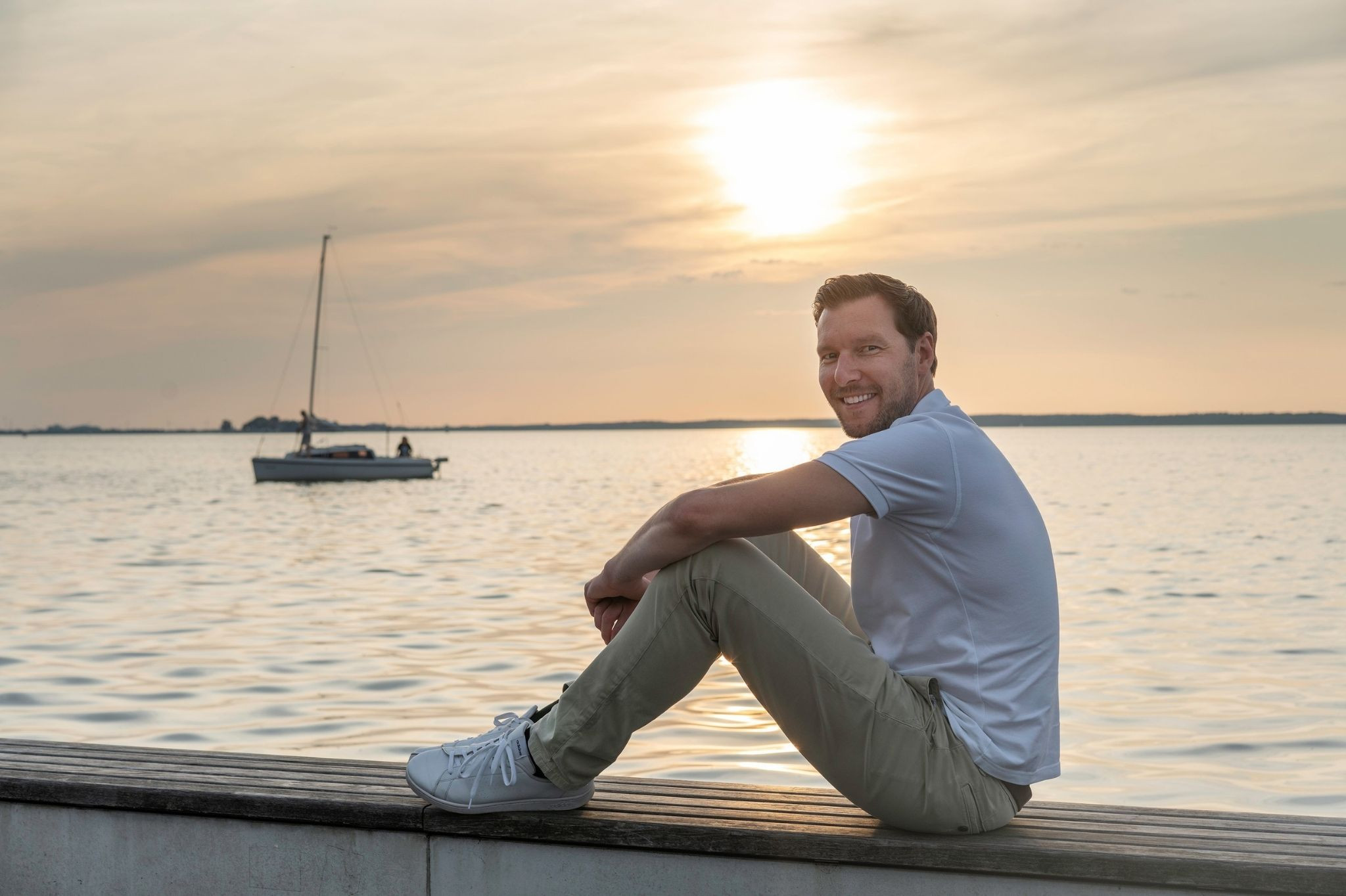 Christian Katz sitzt entspannt am Wasser bei Sonnenuntergang, im Hintergrund ein Segelboot.
