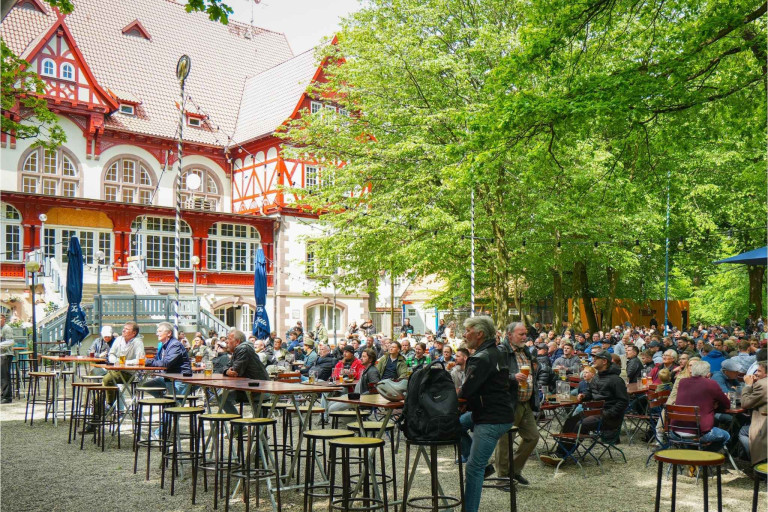 Großer Biergarten mit vielen Menschen vor historischem Gebäude, sonniger Tag, Veranstaltung im Freien