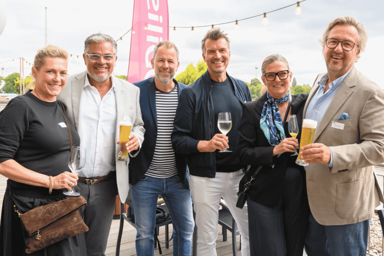 Von links: Dorit und Wolfgang Nestler, Thorsten Baumert, Marco Striewski sowie Sonja und Oliver Bothe. Gruppenfoto von sechs Personen beim nobilis-Sommerfest, mit Getränken in der Hand vor einer pinken Fahne und Lichterkette.