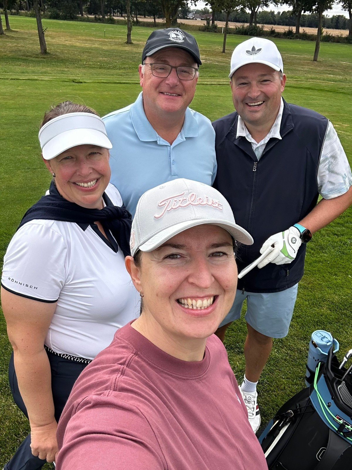 Golfgruppe beim Turnier – 01grasgeflster11ehrgeizverbindet: Vier lachende Menschen auf dem Golfplatz, Teamgeist und Ehrgeiz verbinden sie.