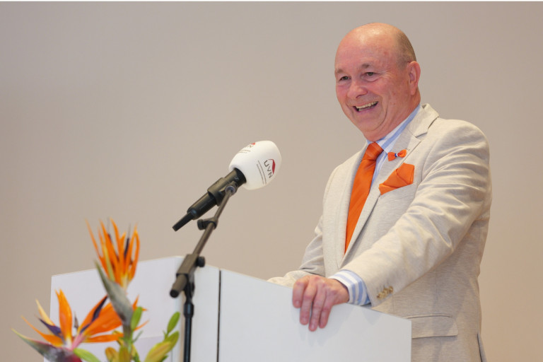 Redner im hellen Anzug und oranger Krawatte am UVN-Mikrofon bei einer Veranstaltung Mann im hellen Anzug mit orangefarbener Krawatte h28lt Rede am Rednerpult mit Mikrofon und UVN-Logo
