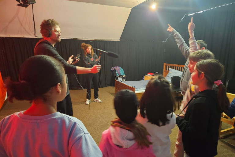Theaterlabor-Workshop mit Kindern und Erwachsenen auf einer Bühne Kinder und Erwachsene beim Theaterlabor-Workshop auf einer Bühne mit Mikrofon und Bett im Hintergrund