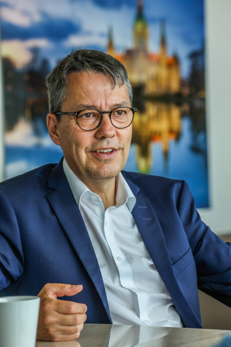 Hansmann im Interview – Porträtfoto eines Mannes im blauen Anzug und weißem Hemd Hansmann im Gespräch, Porträt eines Mannes mit Brille und blauem Anzug