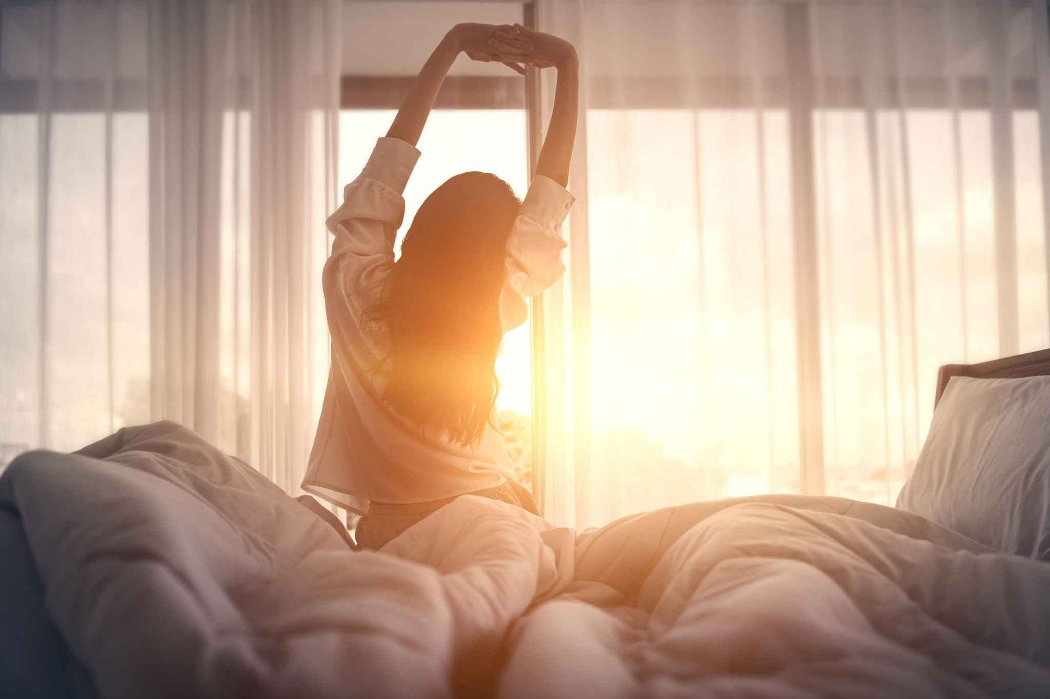 artikel besser schlafen Frau streckt sich morgens im Bett vor Sonnenaufgang