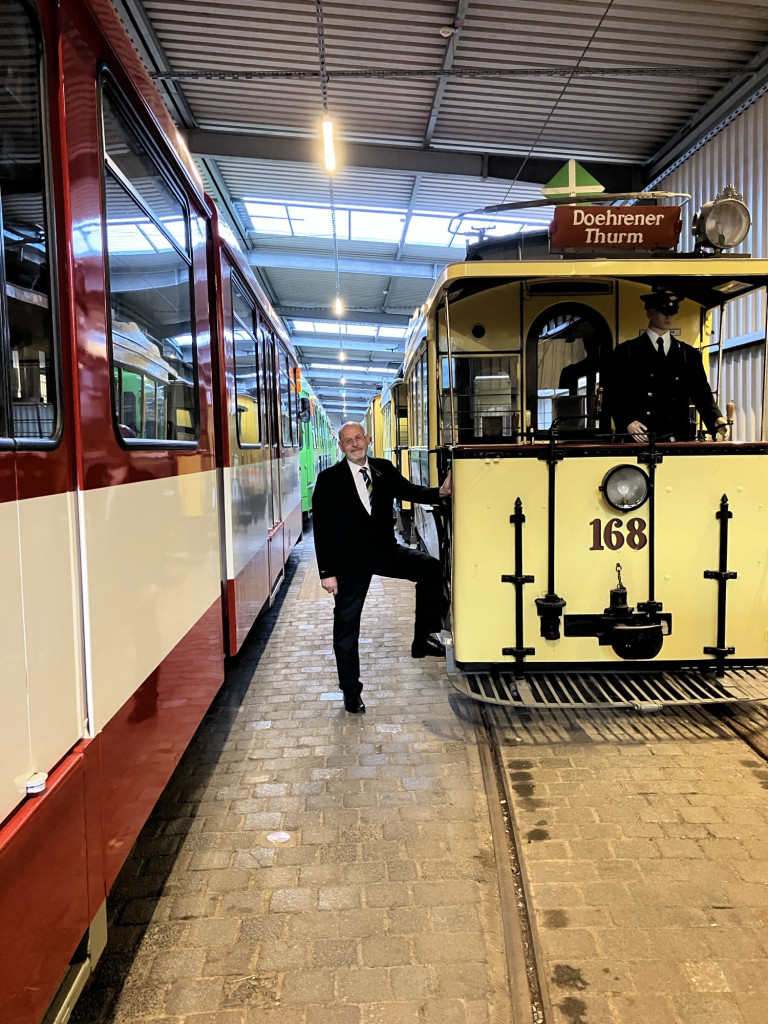 Fritz Faupel bei historischen Straßenbahnen im Straßenbahnmuseum, neben Bahn 168 mit dem Ziel 'Doehrener Thurm'. Fritz Faupel steht zwischen historischen Straßenbahnen, neben einer gelben Bahn mit dem Schild 'Doehrener Thurm'.