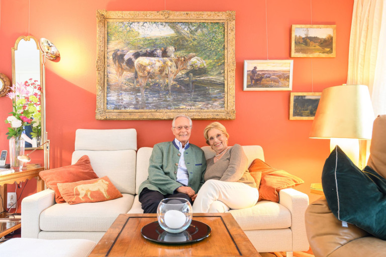 Älteres Paar sitzt glücklich auf dem Sofa, Symbol für lange Liebe, mit Gemälden im Hintergrund