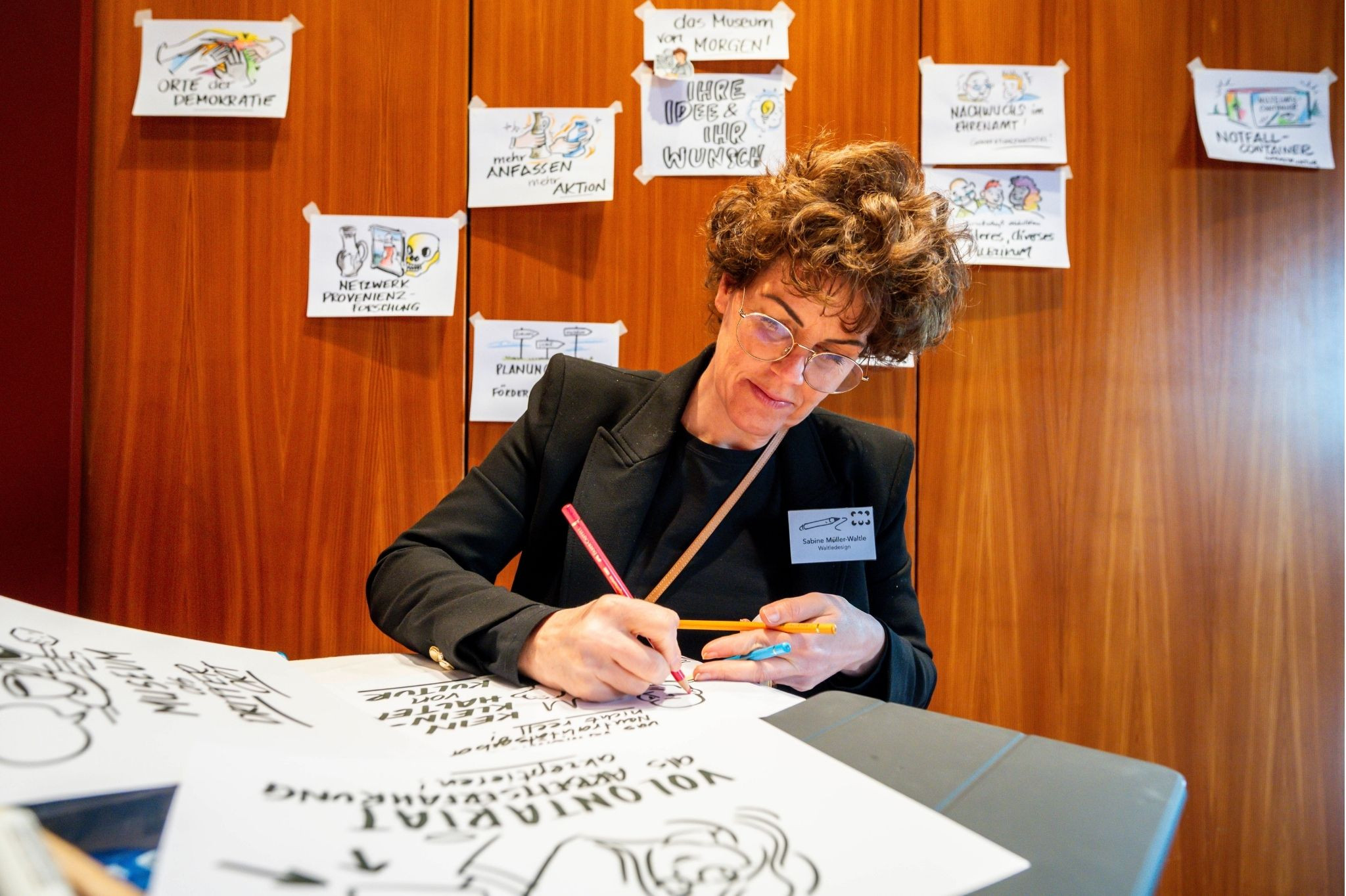 Sabine Müller-Waltle bei einer kreativen Zeichenaktion auf einer Veranstaltung, umgeben von Skizzen und Notizen an der Wand.