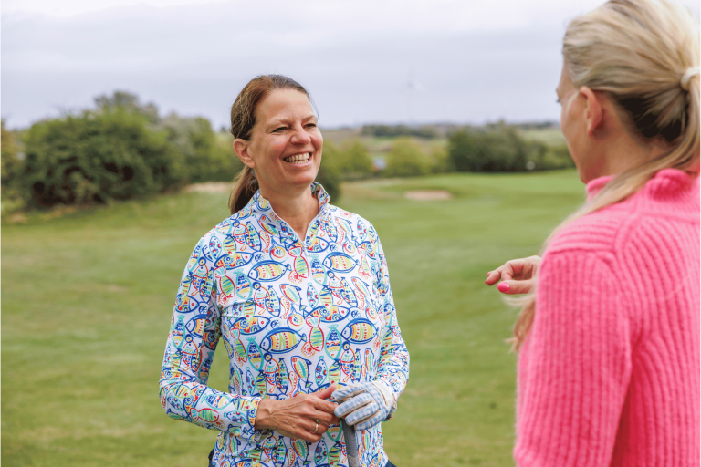 Zwei Frauen unterhalten sich auf dem Golfplatz Nobilis, eine Frau trägt bunte Golfkleidung und lächelt.