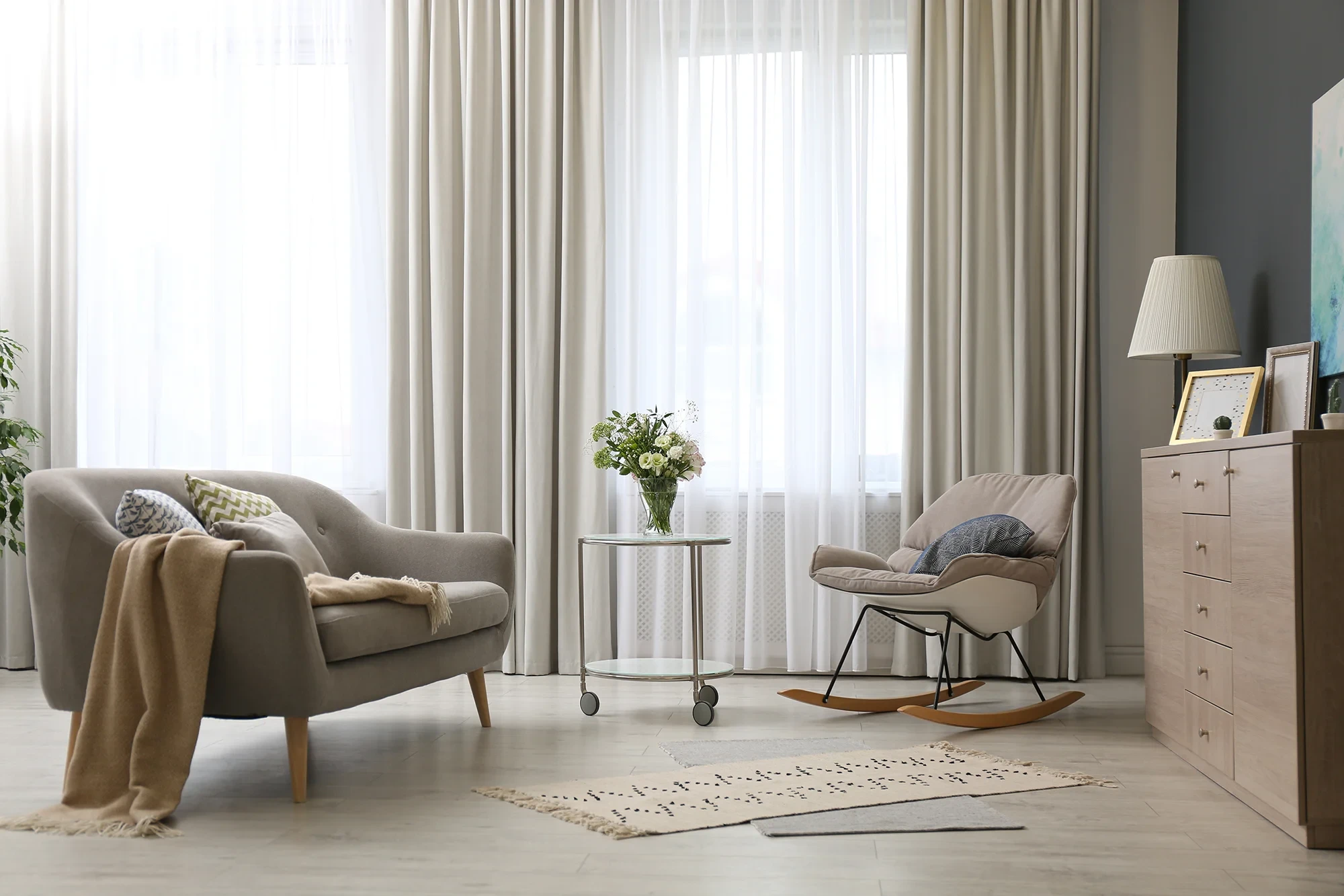 Gemütliches Wohnzimmer als Startseite-Hintergrund mit Sofa und Schaukelstuhl Modernes Wohnzimmer mit Sofa, Schaukelstuhl und Blumenstrauß vor großen Fenstern