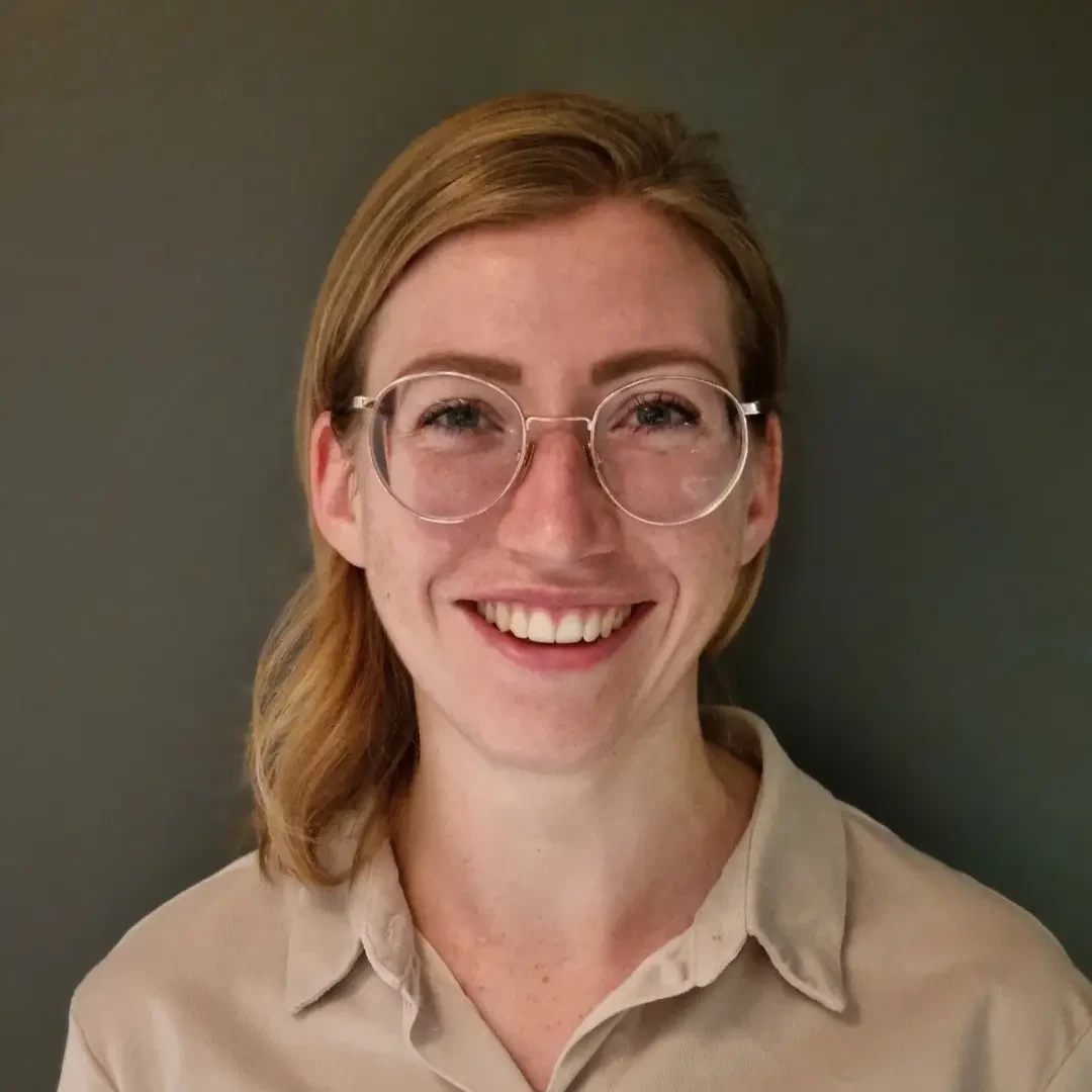Portrait einer lächelnden Frau mit Brille und blonden Haaren vor grauem Hintergrund.