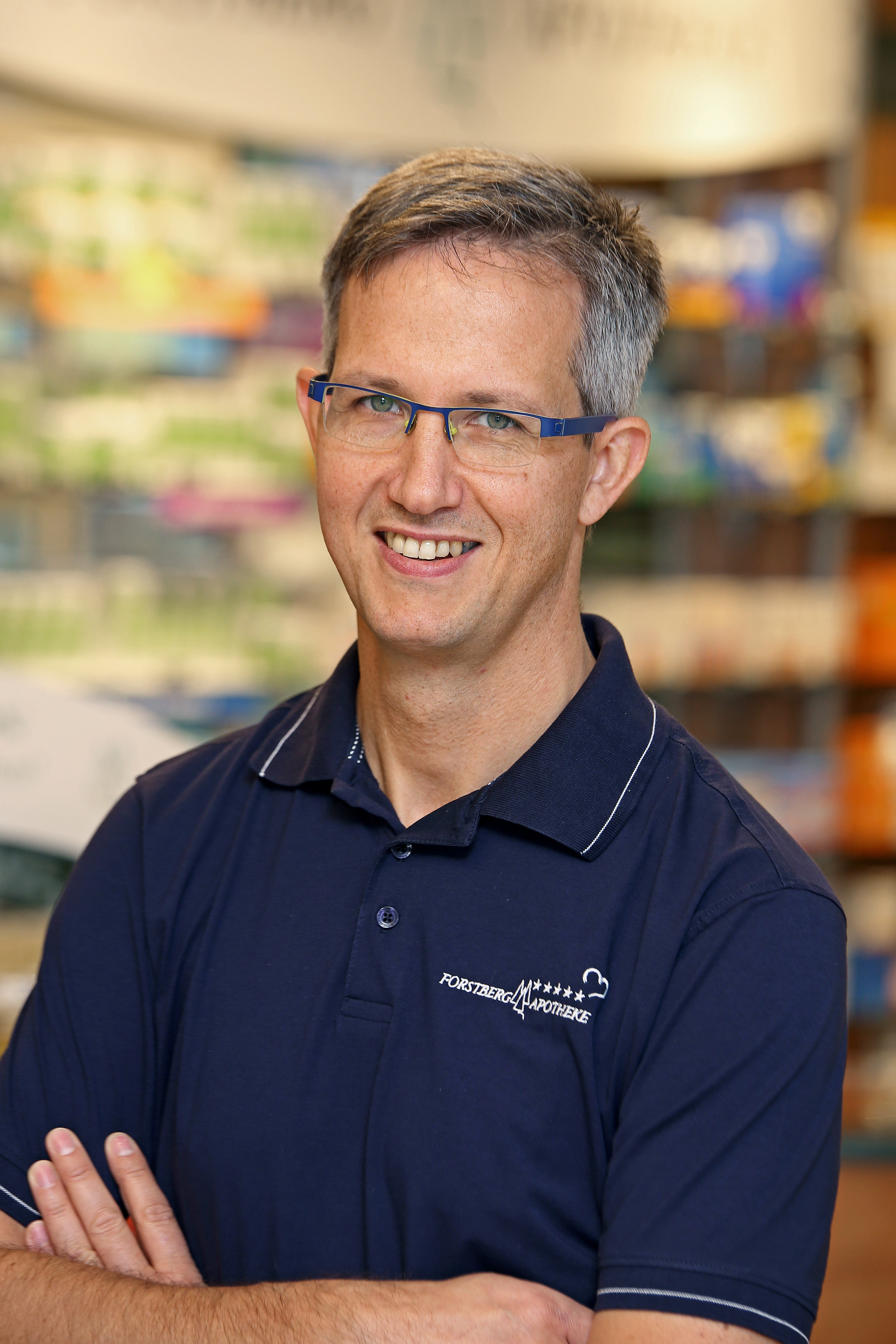 Lächelnder Apotheker mit Brille im blauen Hemd vor Regalen in der Apotheke