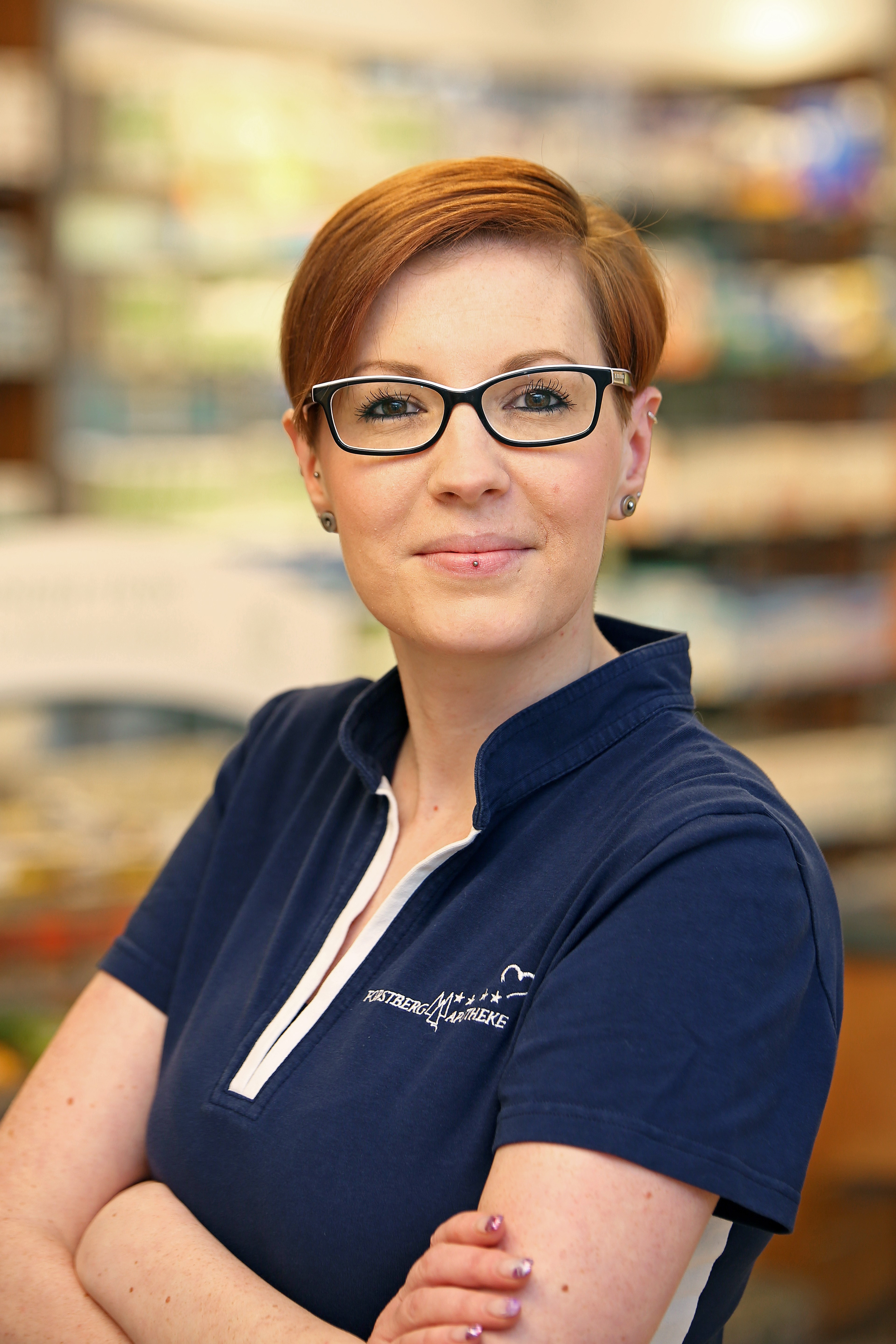 Pharmazeutische Fachkraft in einer Apotheke, trägt eine blaue Dienstkleidung mit Brille.