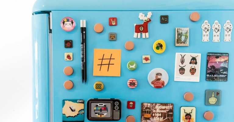 Verschiedene bunte Kühlschrankmagneten auf einer blauen Oberfläche, darunter Motive wie ein Elch, ein Fernseher, ein Hashtag und verschiedene Symbole.