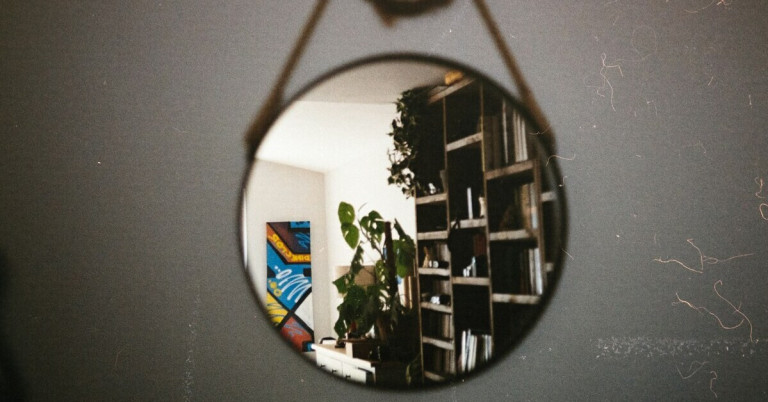 Runder Spiegel an der Wand reflektiert ein Wohnzimmer mit Bücherregal, Pflanzen und bunter Wandkunst