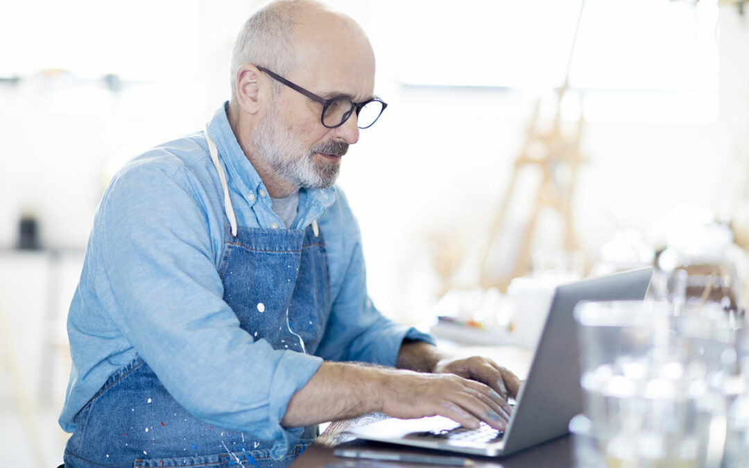 Älterer Mann arbeitet an einem Laptop, trägt eine blaue Schürze