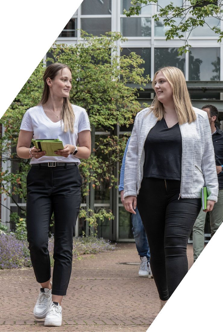 Zwei junge Frauen gehen mit Notizblöcken auf einem Campus entlang, moderne Kleidung, Frühlingswetter