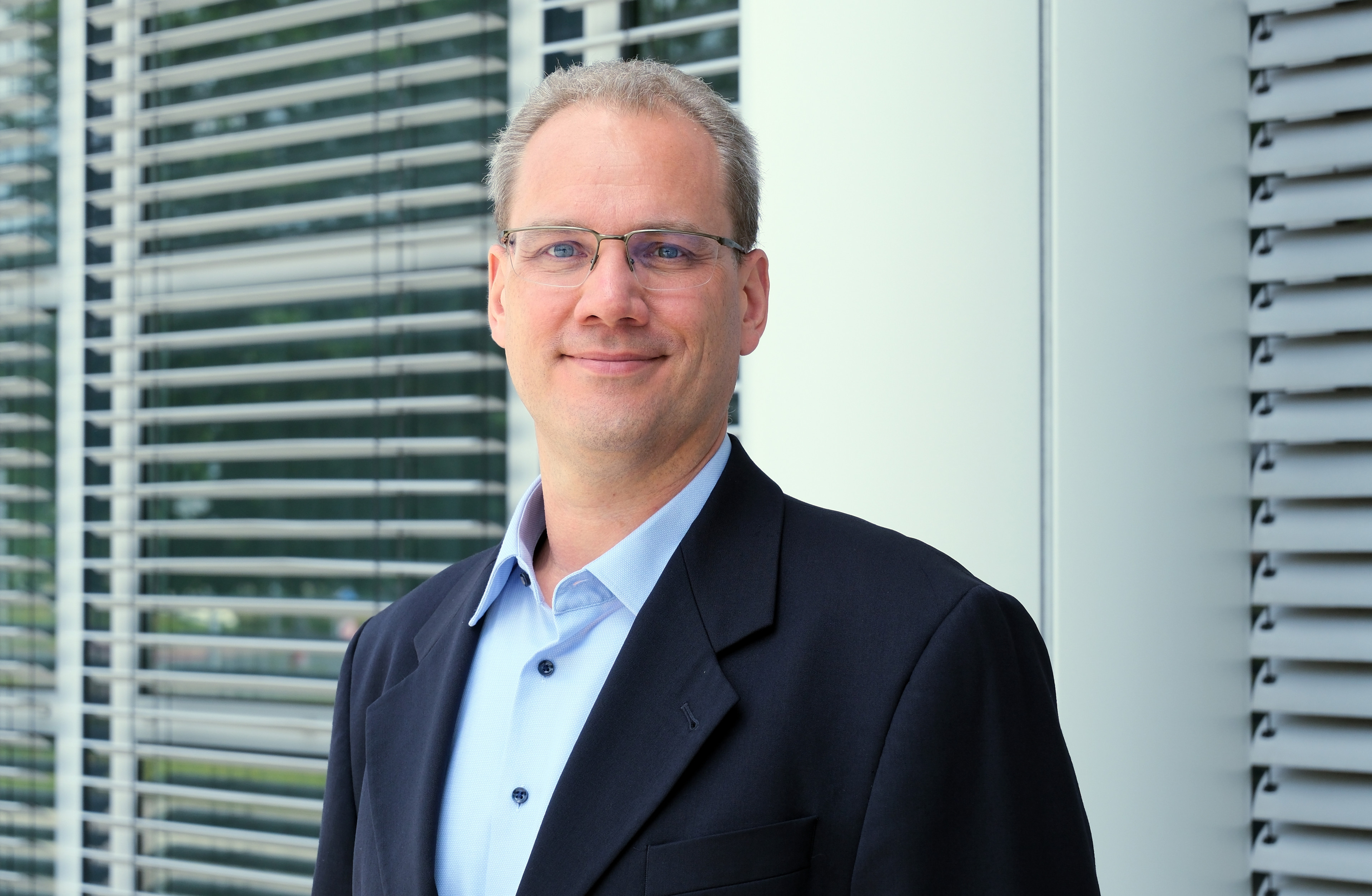 Porträt von Thomas Hortig im Querformat, Mann mit Brille und Anzug vor modernem Hintergrund