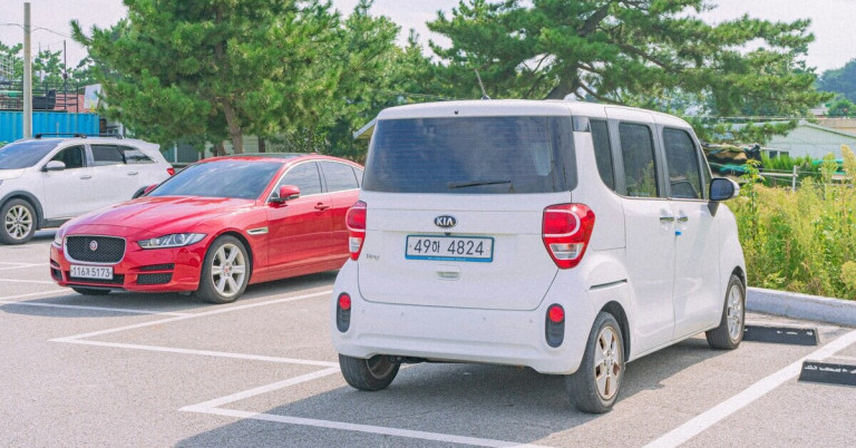 Weißes Auto auf Parkplatz – Kia Fahrzeug im Sommer neben rotem Auto Weißes Auto auf Parkplatz, Kia Fahrzeug neben rotem Auto, Sommer, Bäume im Hintergrund