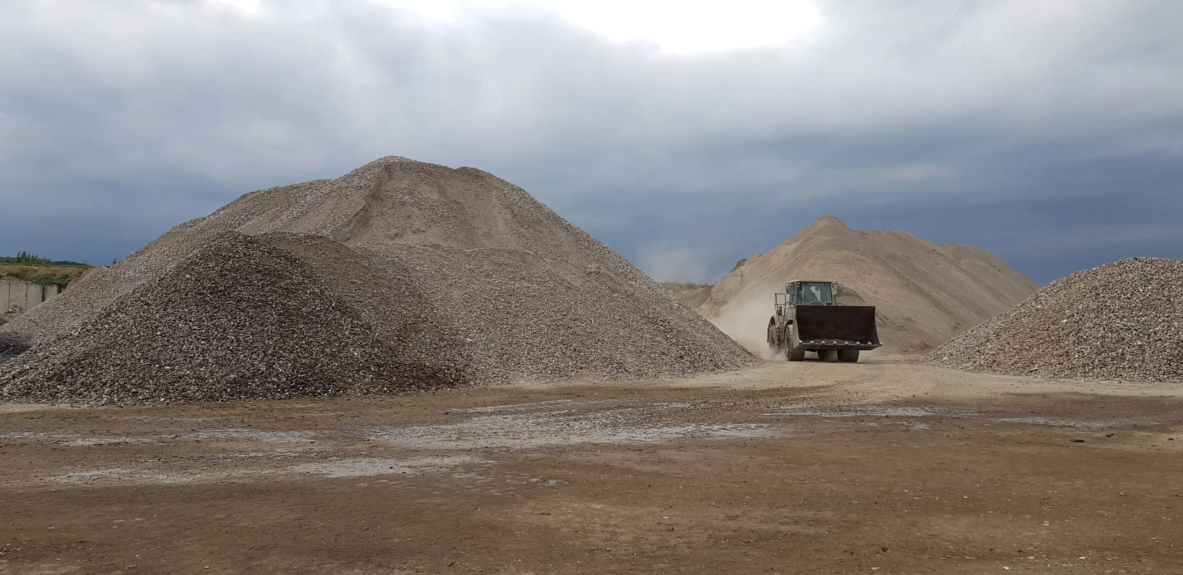 Radlader bewegt Kies auf einem Schotterplatz mit großen Kieshaufen unter bewölktem Himmel