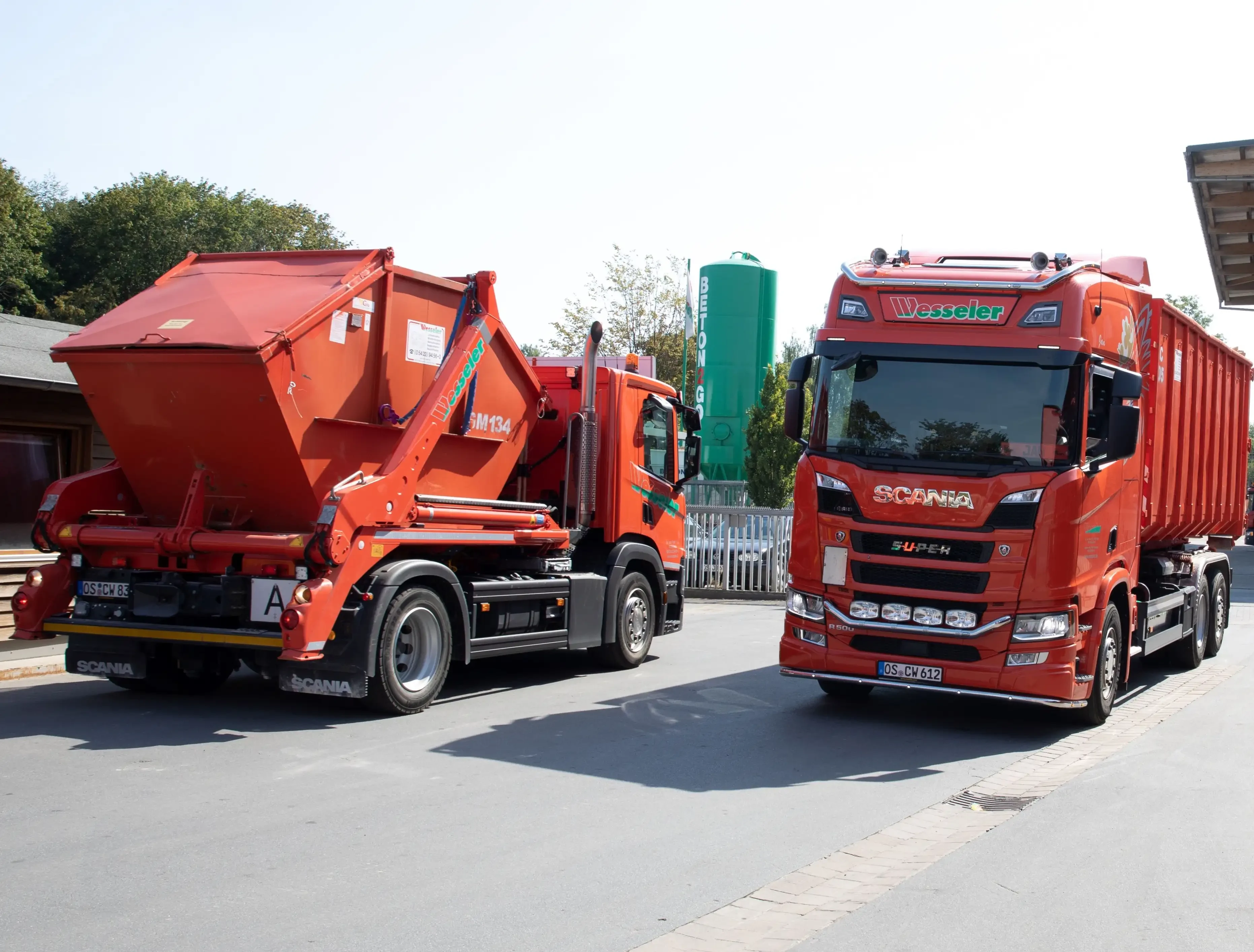 Zwei rote LKWs der Firma Wesseler auf einem Betriebsgelände, einer davon ein Scania mit Containeraufbau.