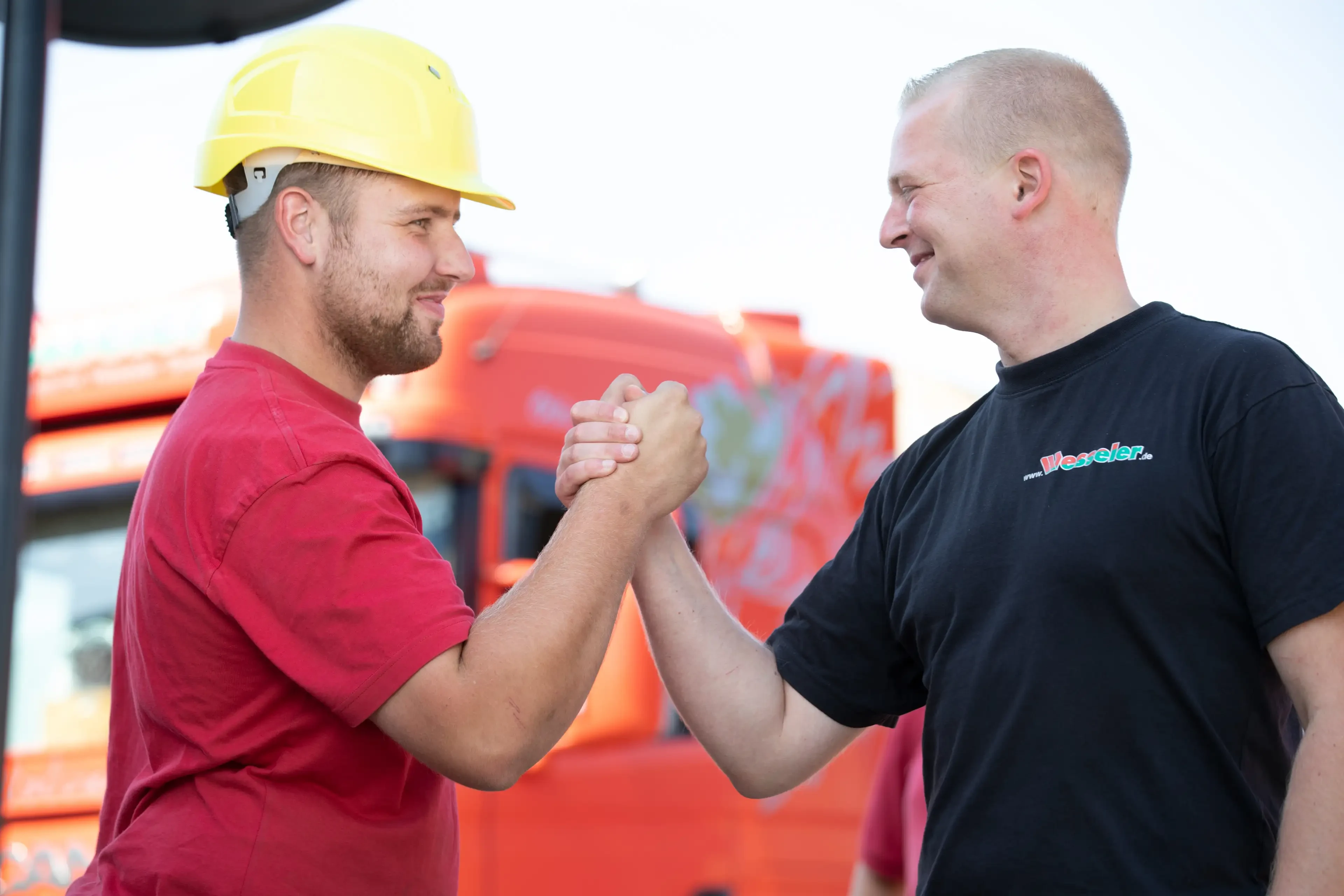 Zwei Männer geben sich einen festen Händedruck vor einem roten LKW, einer trägt einen Bauhelm