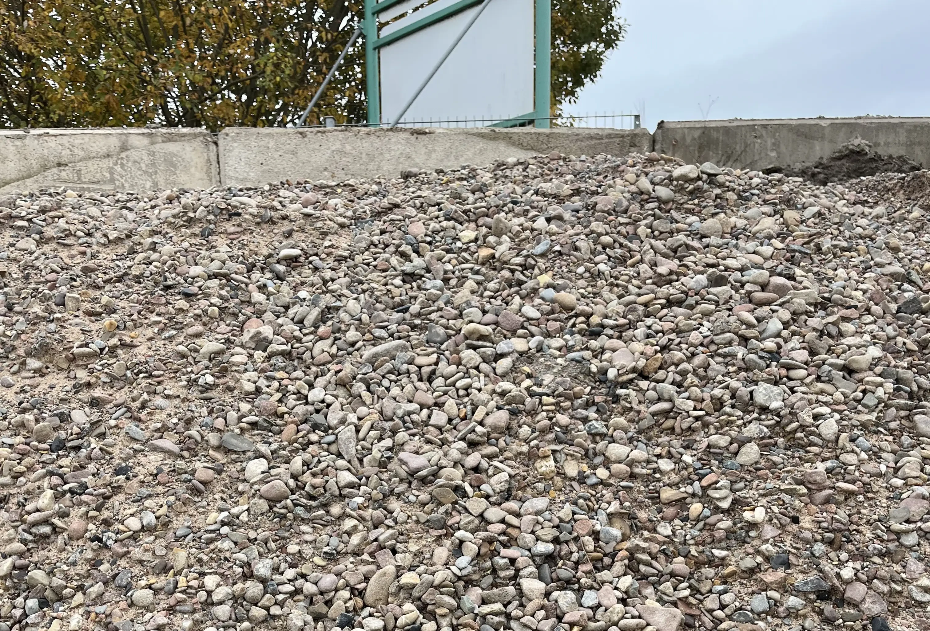 Kieshaufen auf einem Bauhof mit Betonmauer und Bäumen im Hintergrund