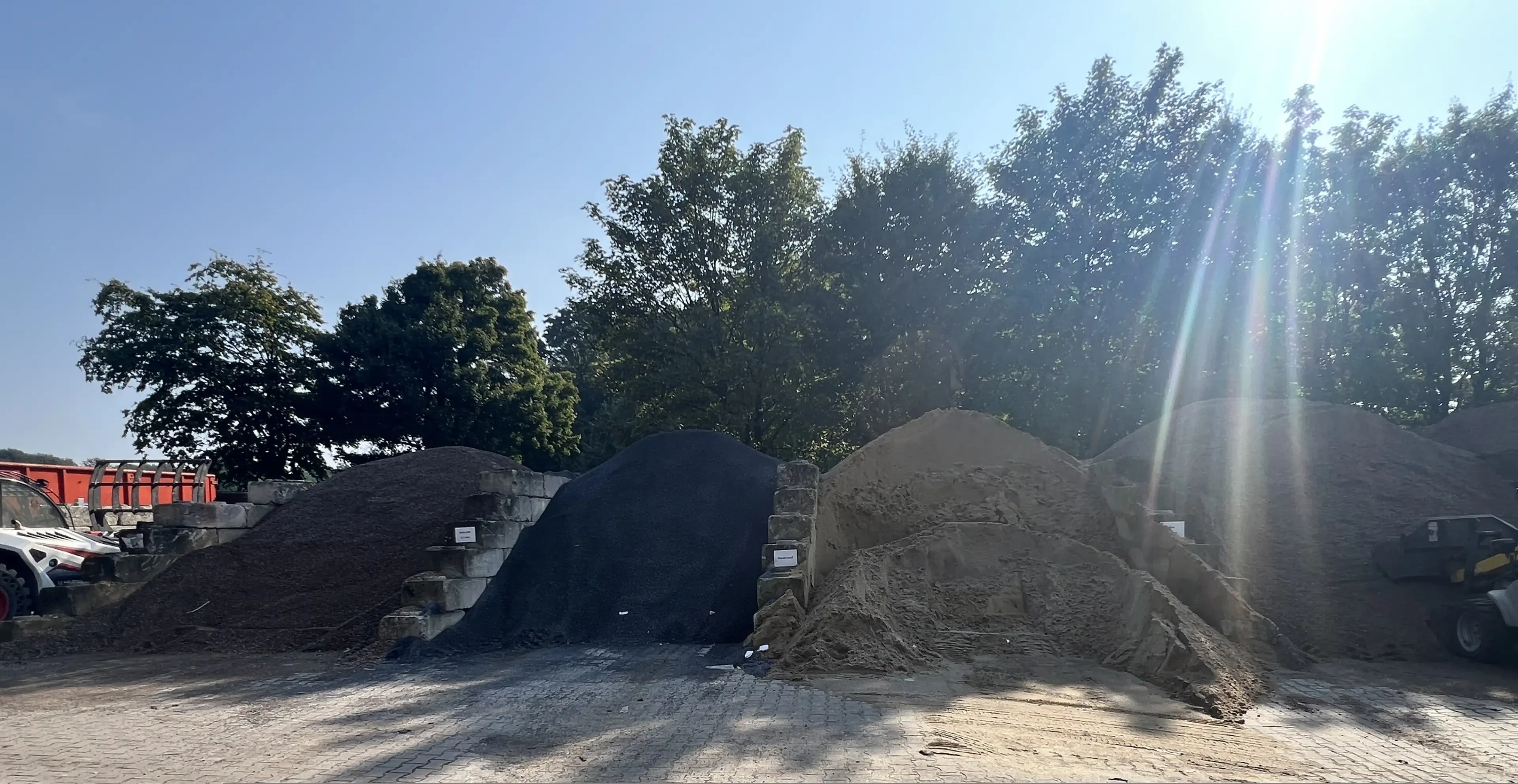 Verschiedene Schüttgüter wie Kies und Sand vor einer Baumreihe auf einem Lagerplatz