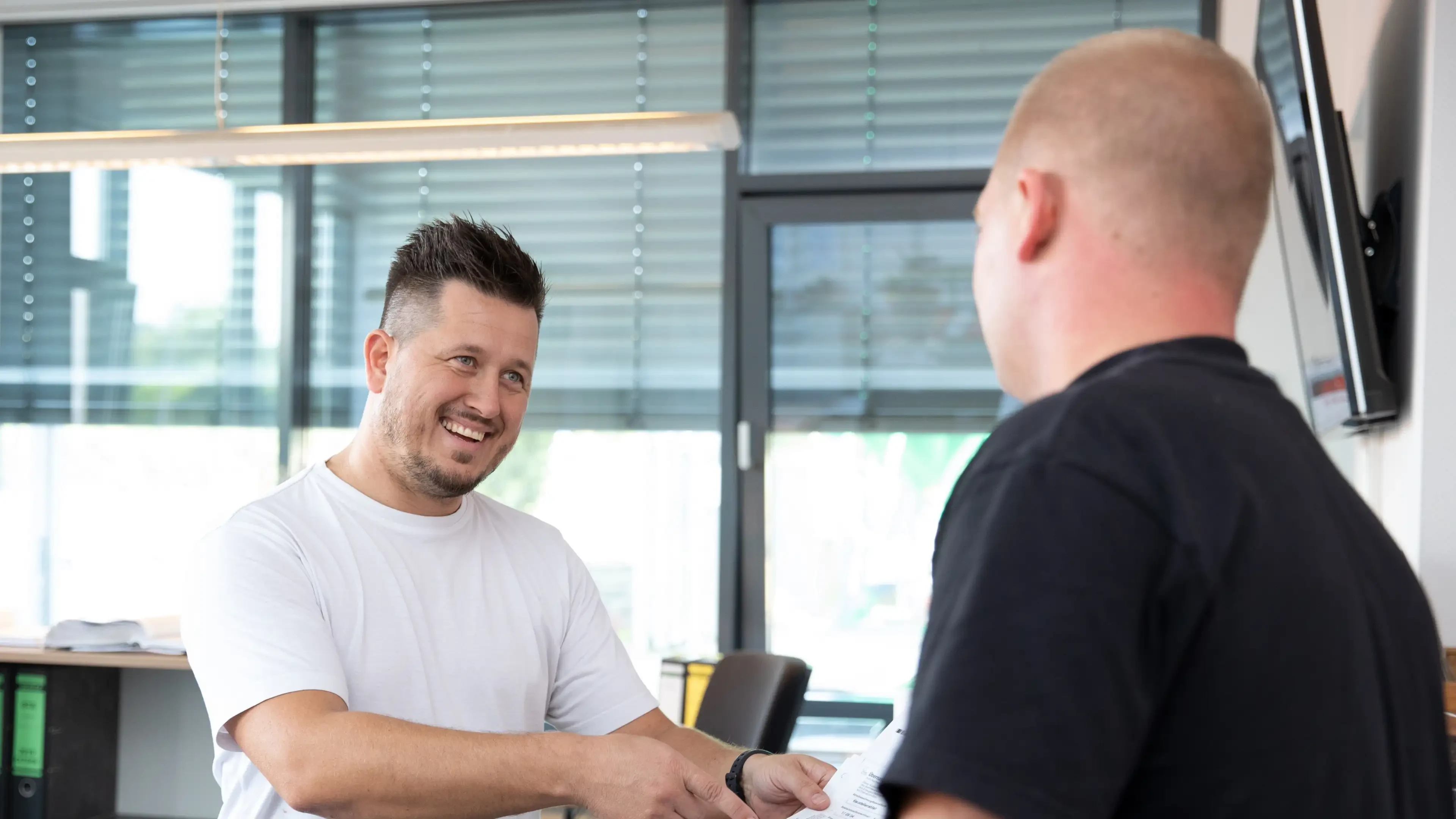 Zwei Männer im Gespräch in einem modernen Büro, einer übergibt dem anderen ein Dokument