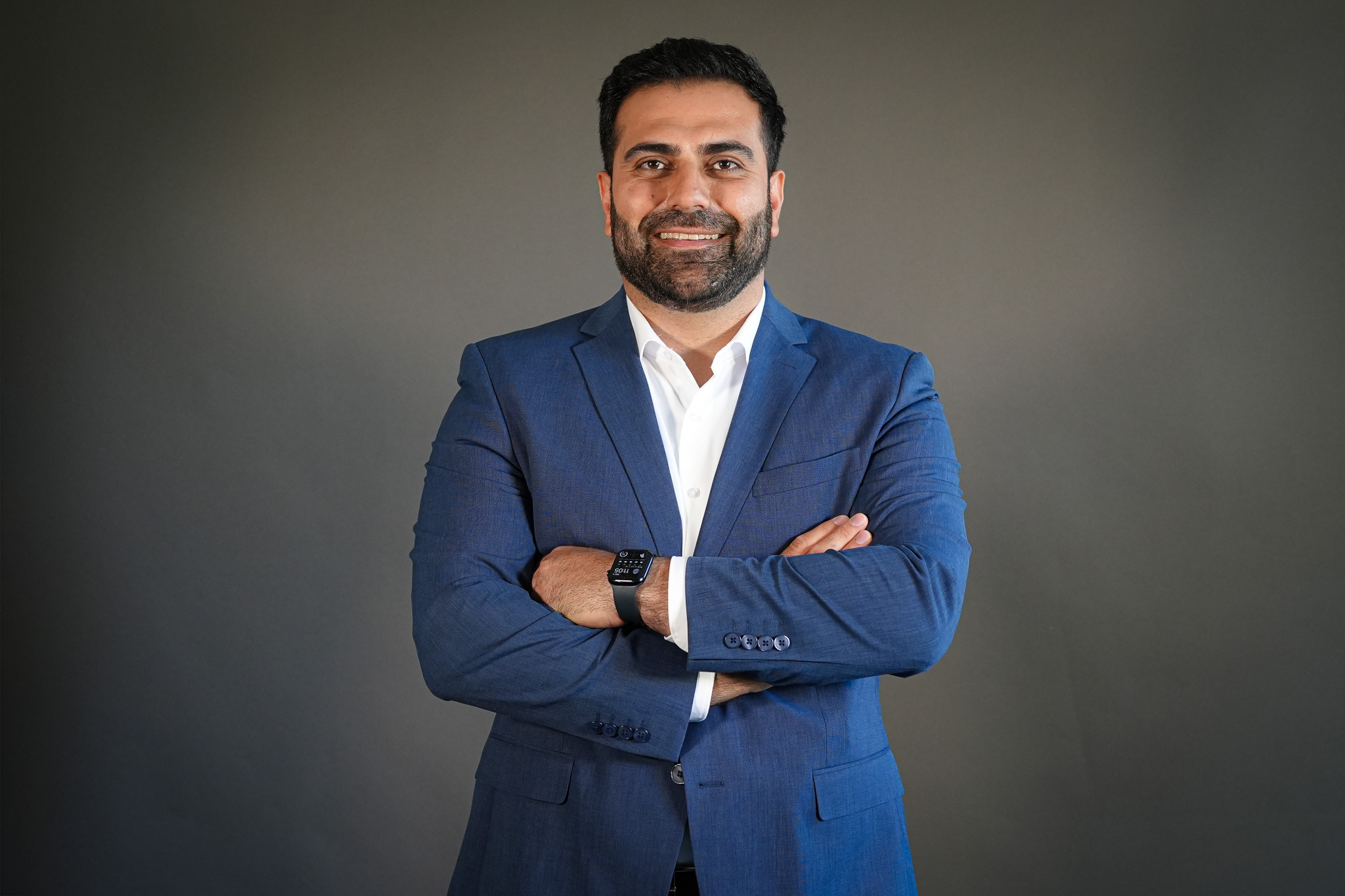 Milad Shams im Business-Outfit mit verschränkten Armen Milad Shams im blauen Anzug mit verschränkten Armen vor grauem Hintergrund