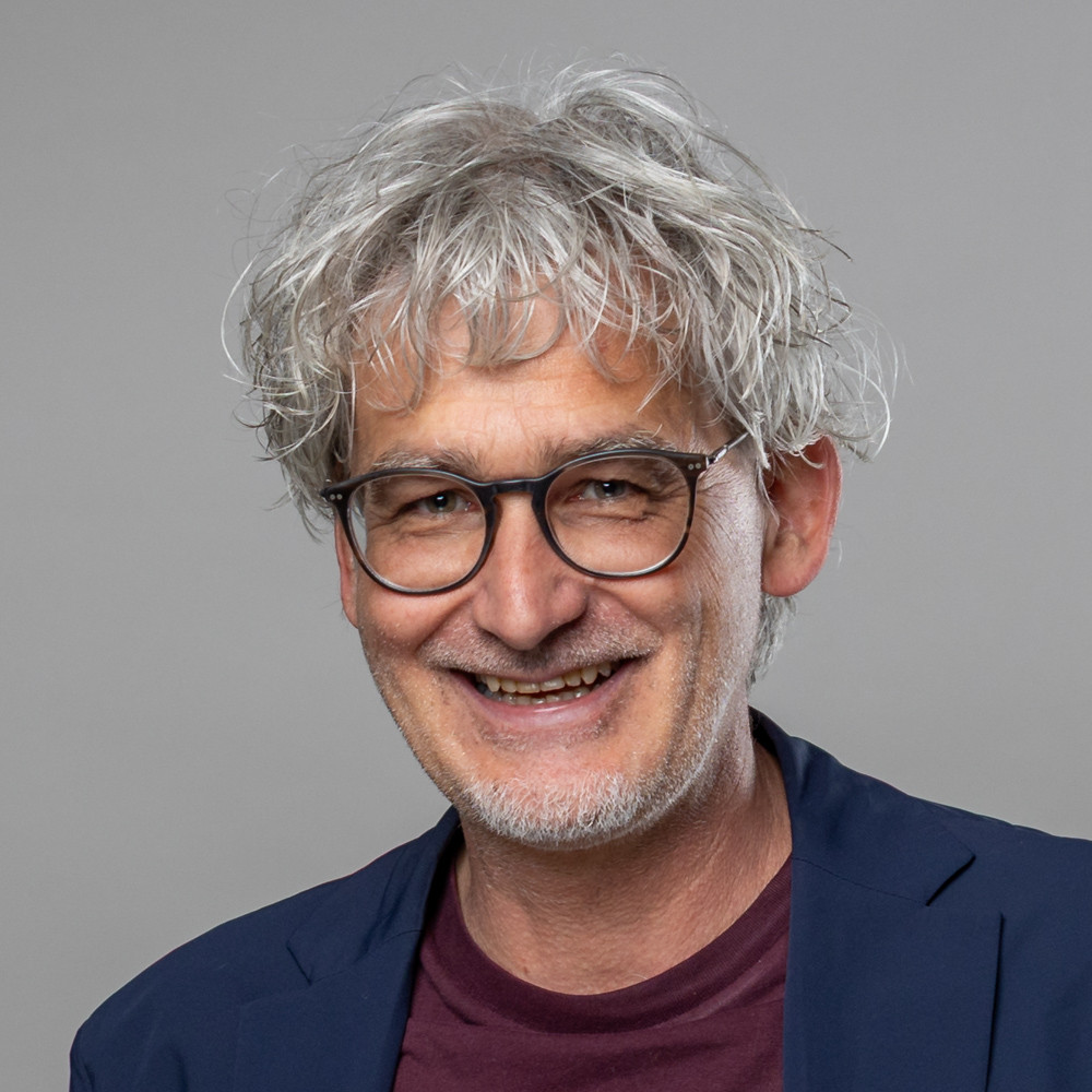 Porträt von Andi Stuckl mit grauen Haaren und blauem Sakko Andi Stuckl mit grauen Haaren und blauem Sakko vor grauem Hintergrund