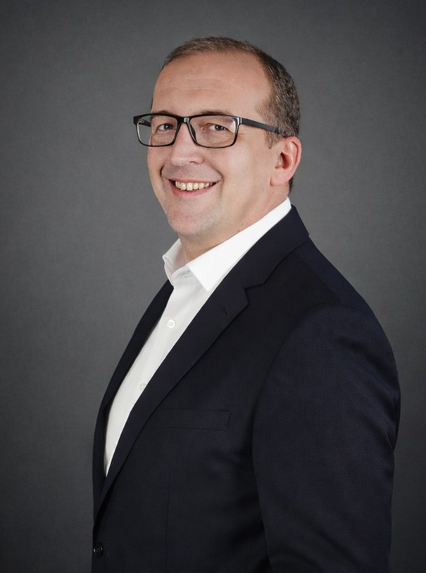 Profilbild eines lächelnden Mannes mit Brille im Anzug vor grauem Hintergrund