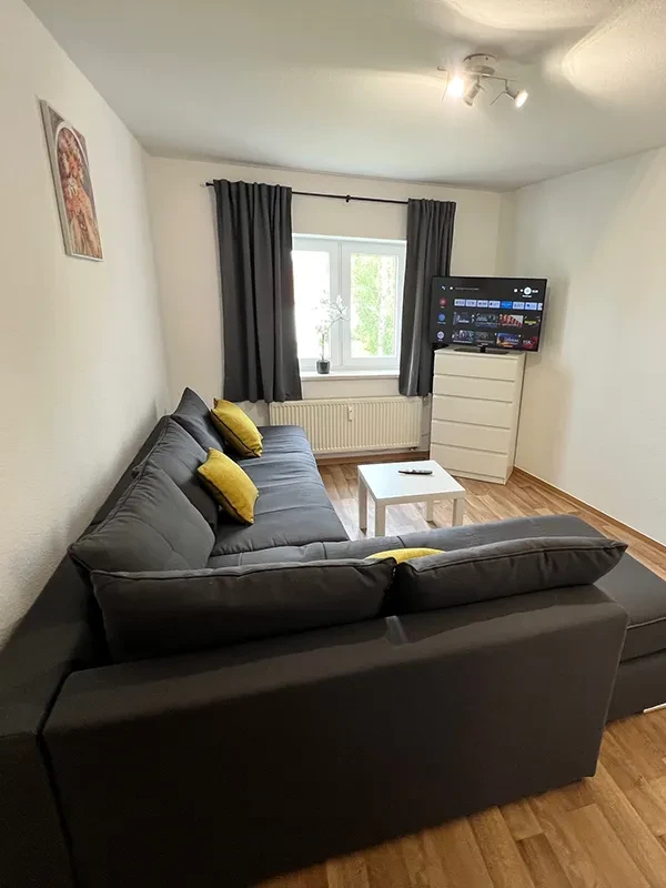 Großes Apartment Wohnzimmer – modernes Interieur mit Sofa, Fernseher und Fenster Modernes großes Apartment Wohnzimmer mit grauem Sofa, gelben Kissen, TV und Fenster