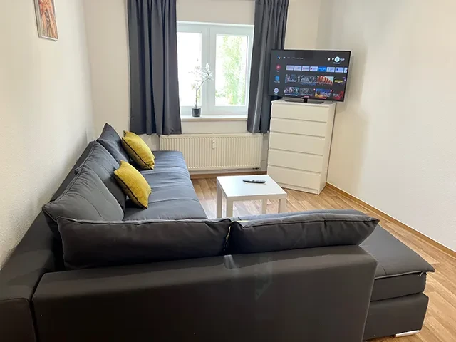Apartment groß – Modernes Wohnzimmer mit Sofa, Fernseher und Fenster Modernes Wohnzimmer in einem großen Apartment mit grauem Sofa, gelben Kissen, Fernseher und Fenster
