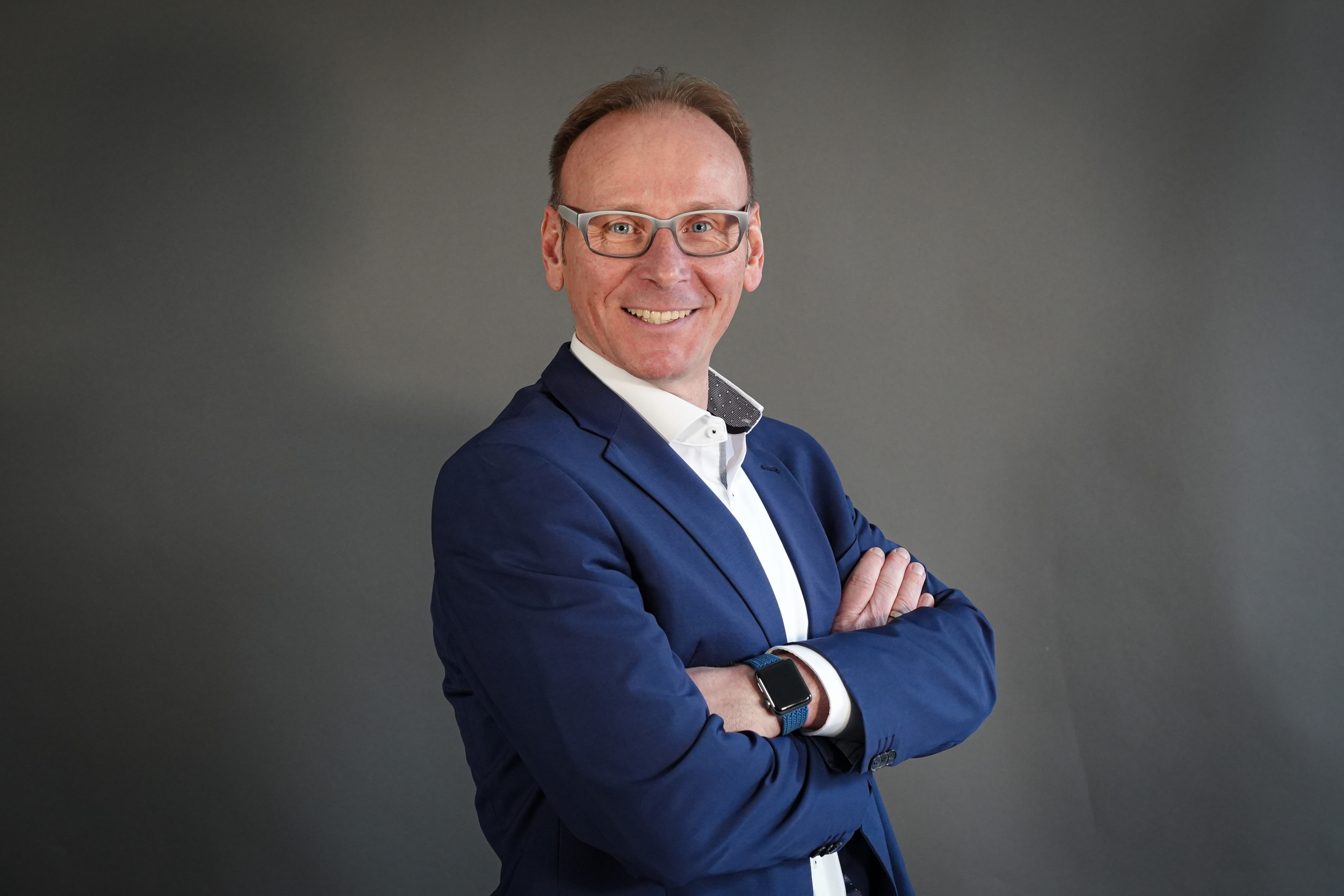 Bert König im blauen Anzug mit verschränkten Armen vor grauem Hintergrund