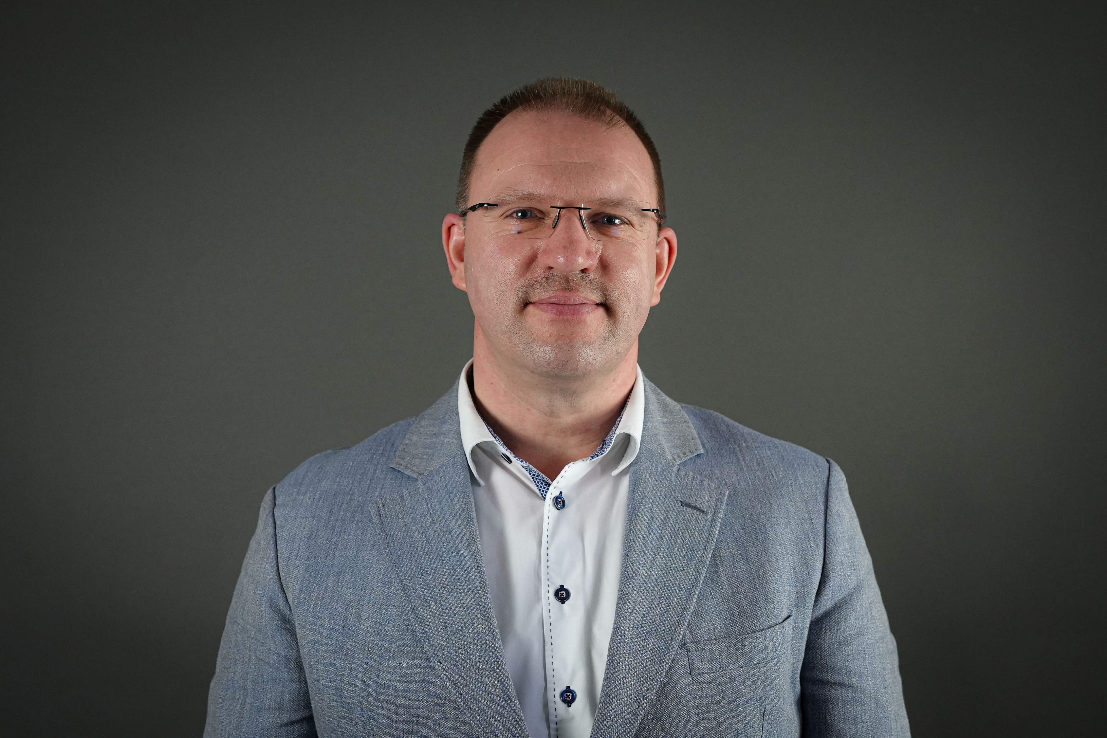 Thomas im klassischen Business-Outfit mit grauem Sakko und weißem Hemd vor neutralem Hintergrund