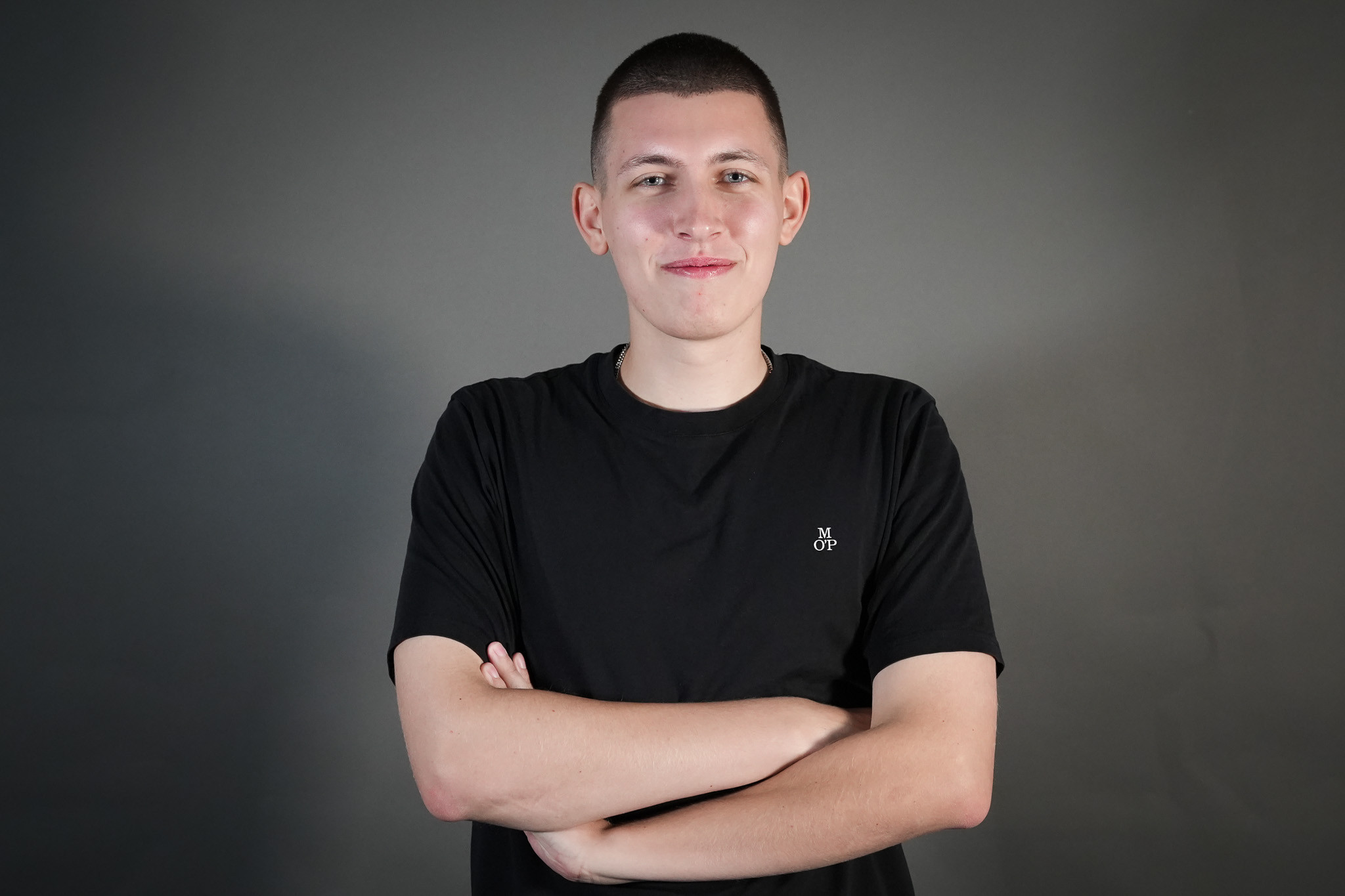 Person mit schwarzem T-Shirt mit MOP-Logo vor grauem Hintergrund mit verschränkten Armen