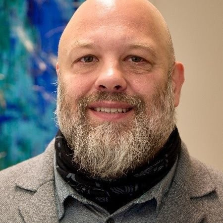 Mann mit grauem Bart und grauem Sakko vor abstraktem Hintergrund