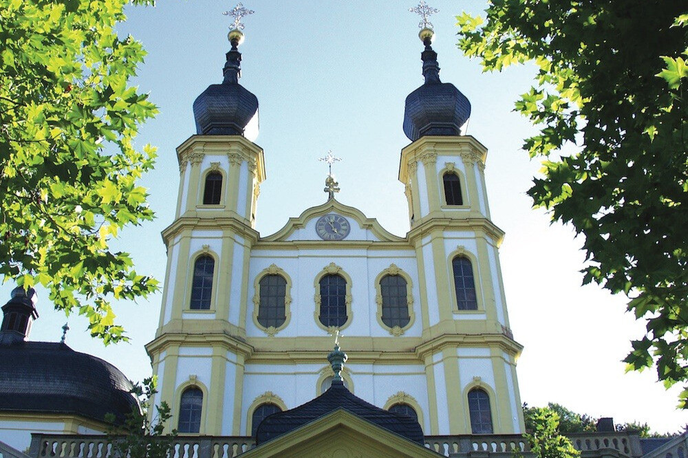 Barocke Kirche mit zwei Türmen und Uhr an der Fassade, im Sonnenschein, umgeben von Bäumen