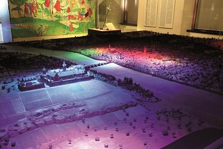Beleuchtetes Stadtmodell in einem Museum mit farbigen Lichtern und mittelalterlicher Wandmalerei im Hintergrund