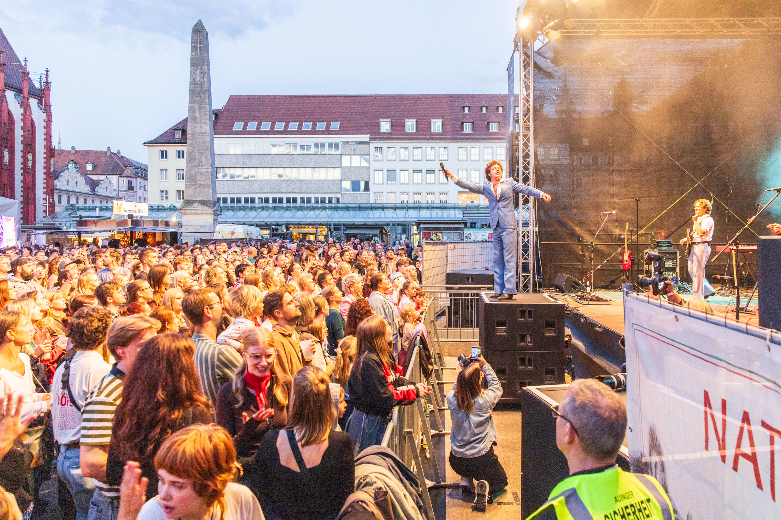 Open-Air-Konzert mit großer Menschenmenge vor einer Bühne am Abend, Künstler auf der Bühne animiert das Publikum, Stadtplatz mit Obelisk im Hintergrund