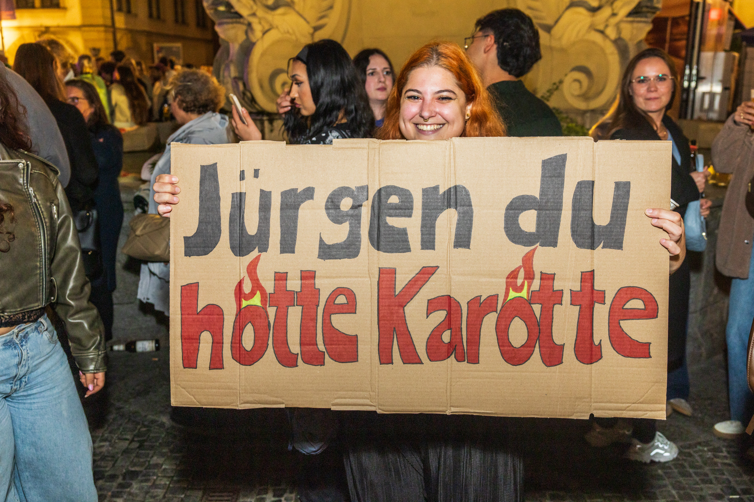 Frau hält ein Pappschild mit der Aufschrift 'Jürgen du hötte Karötte' auf einer Veranstaltung