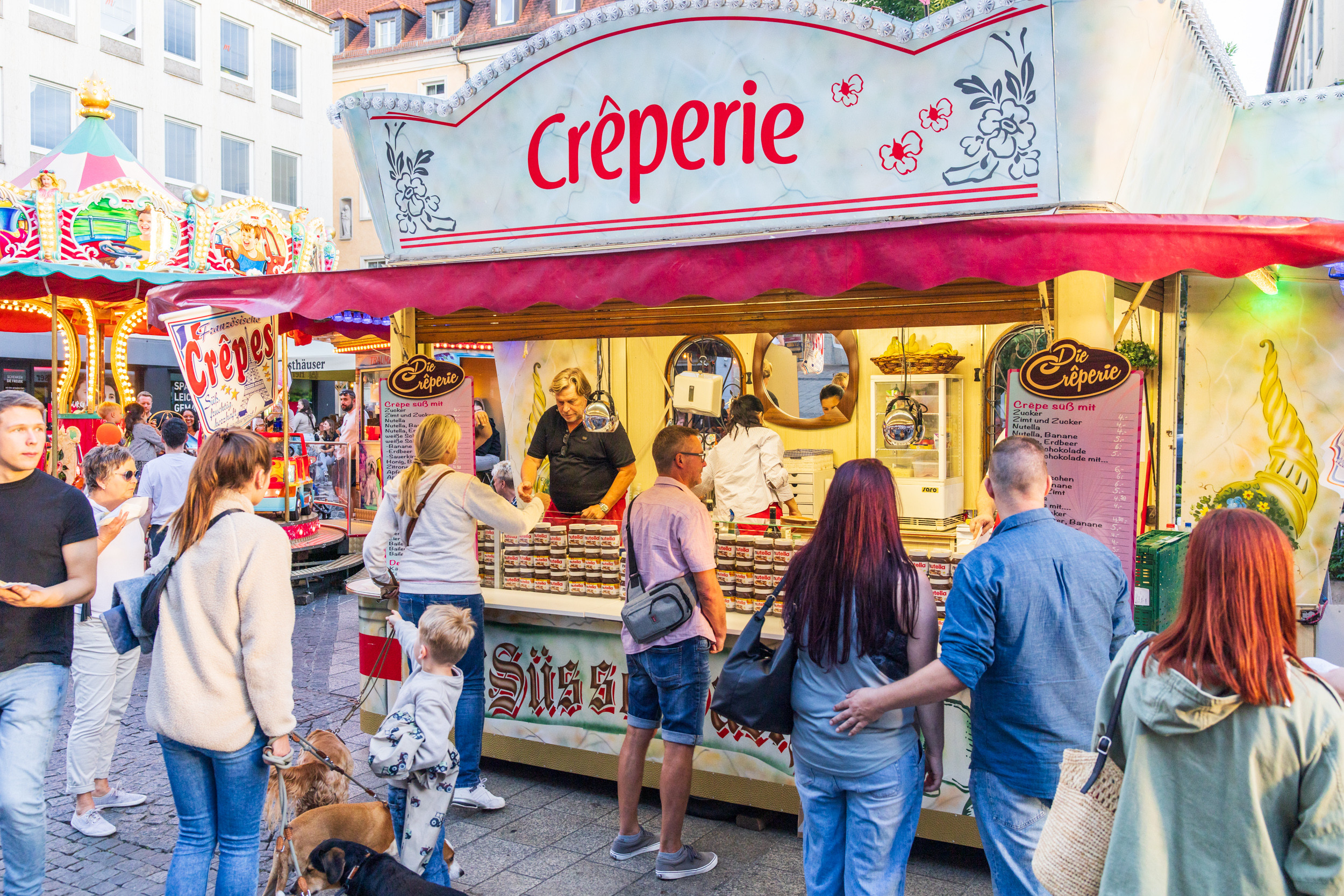Menschen stehen an einer Crêperie auf einem Stadtfest und bestellen Crêpes.