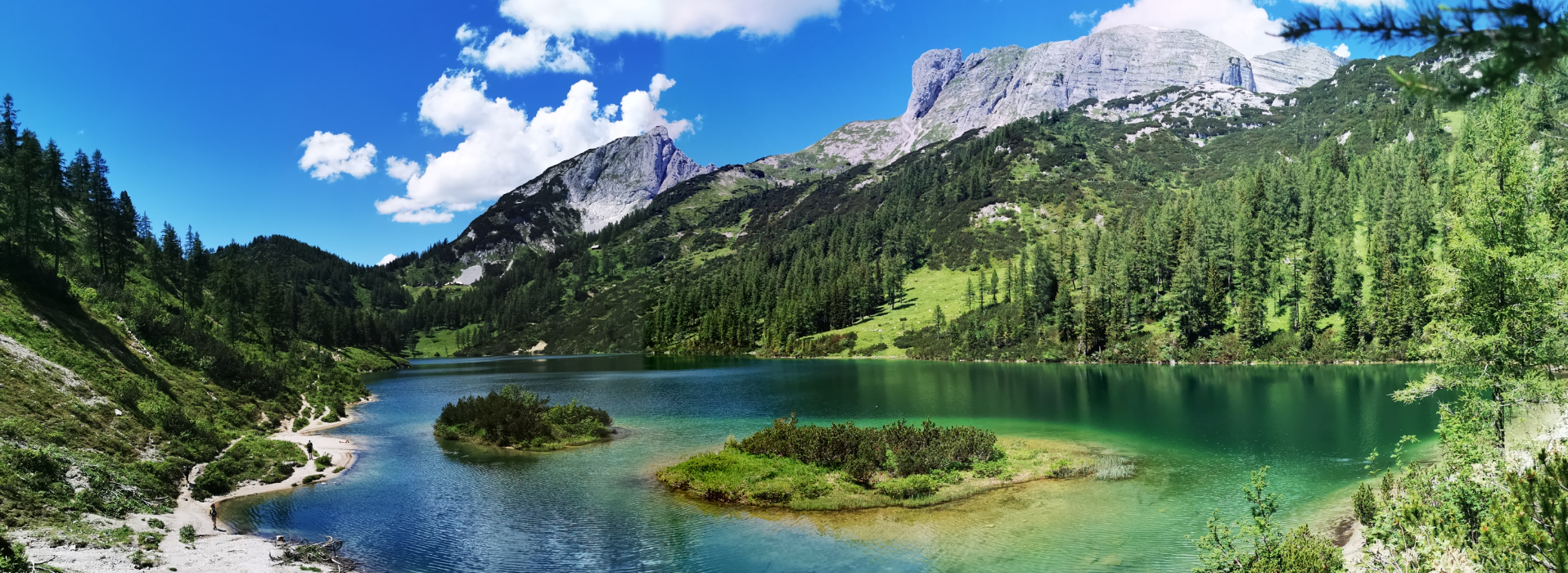 Bergsee mit klar blauem Wasser, umgeben von grünen Wäldern und hohen Bergen unter blauem Himmel