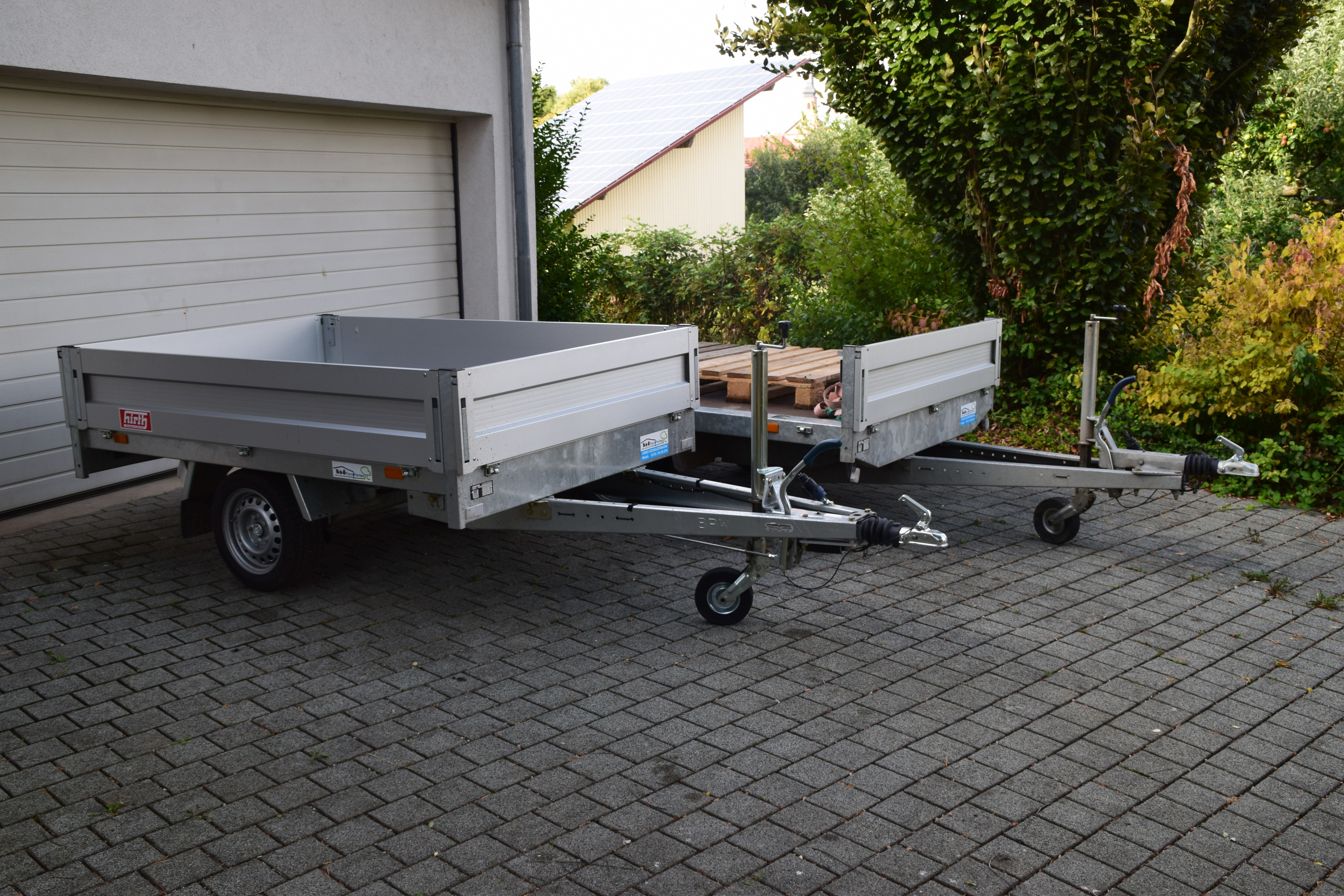 Zwei PKW-Anhänger auf einem gepflasterten Hof vor einer Garage