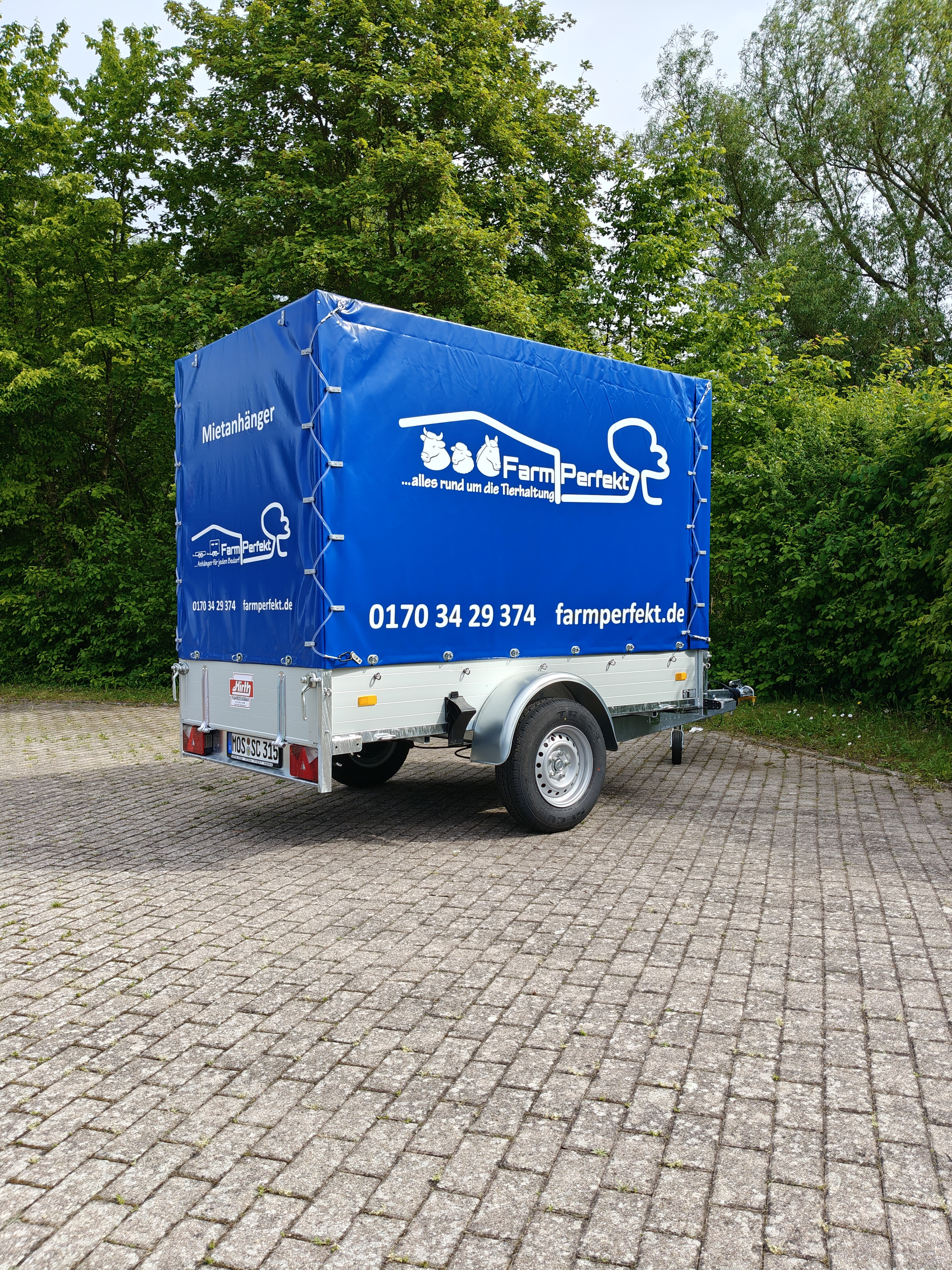 Mietanhänger von FarmPerfekt mit blauer Plane auf Parkplatz, Werbung für Tierhaltung und Anhängervermietung