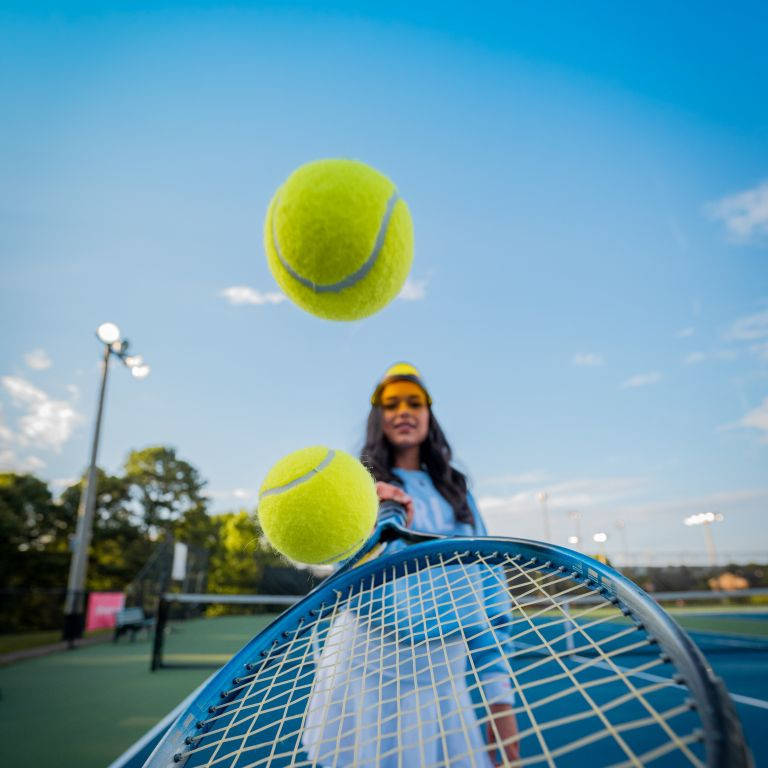 Tennisspielerin auf einem Tennisplatz mit Tennisschläger und Tennisbällen im Vordergrund bei blauem Himmel