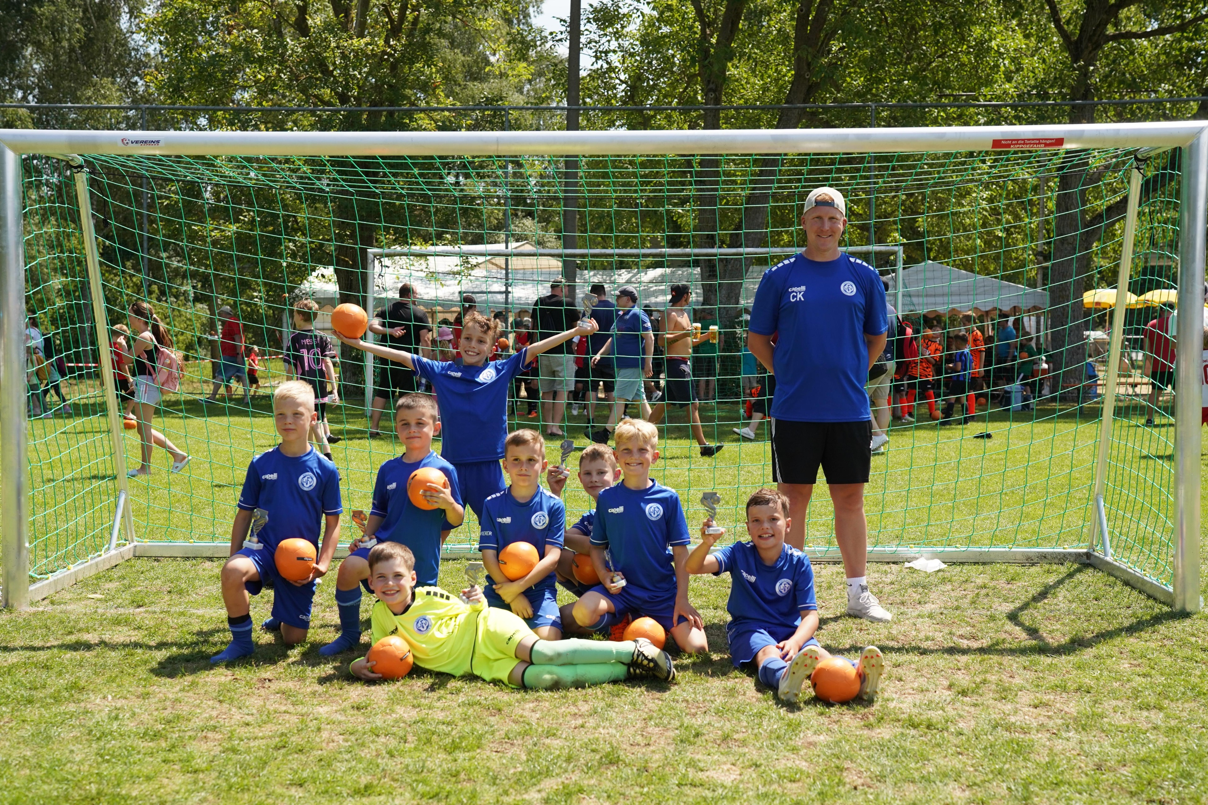 Junges Fußballteam posiert mit ihrem Trainer vor einem Tor auf einem Sportplatz, halten Pokale und Bälle.