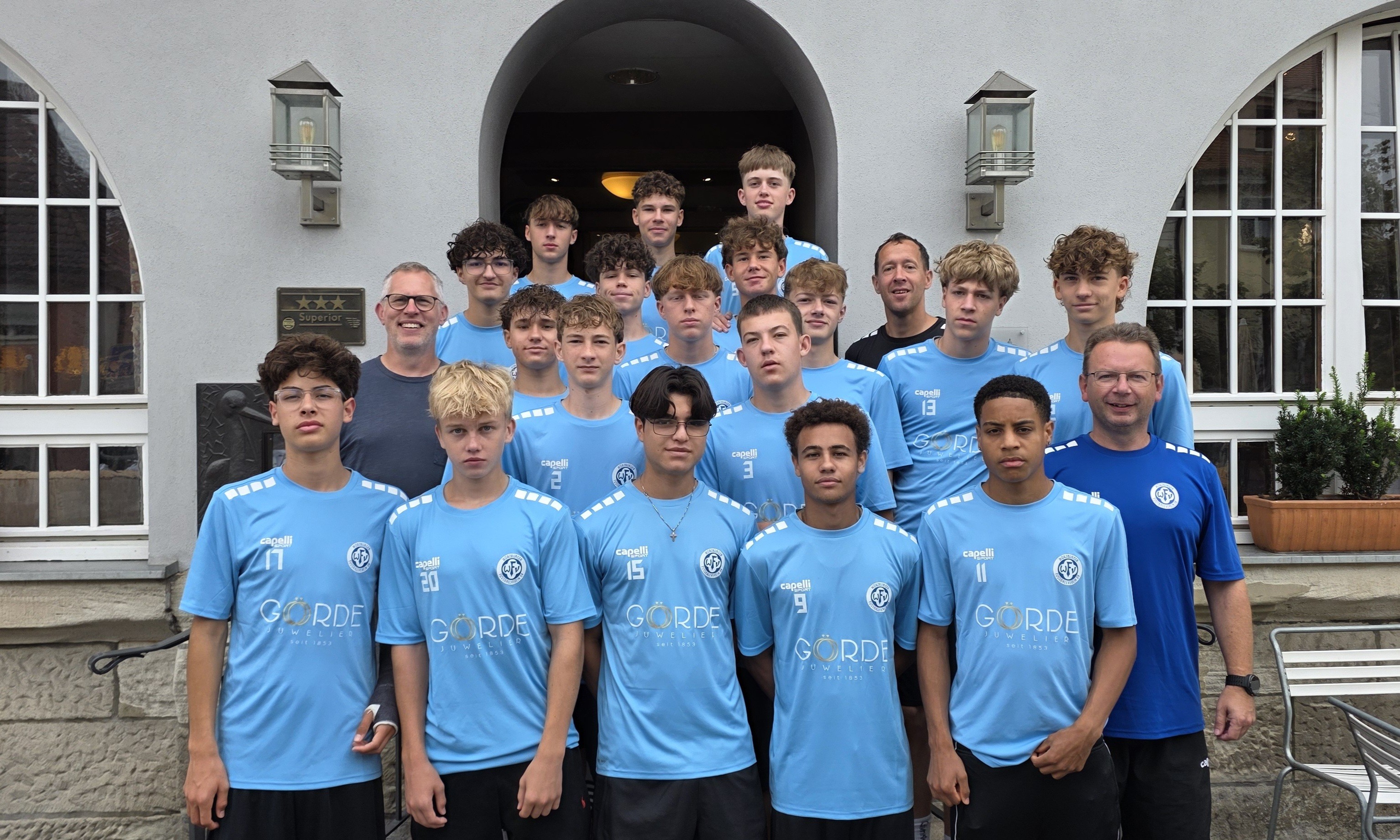 Jugend-Fußballmannschaft in hellblauen Trikots mit Sponsorenlogo Görde vor einem Hoteleingang