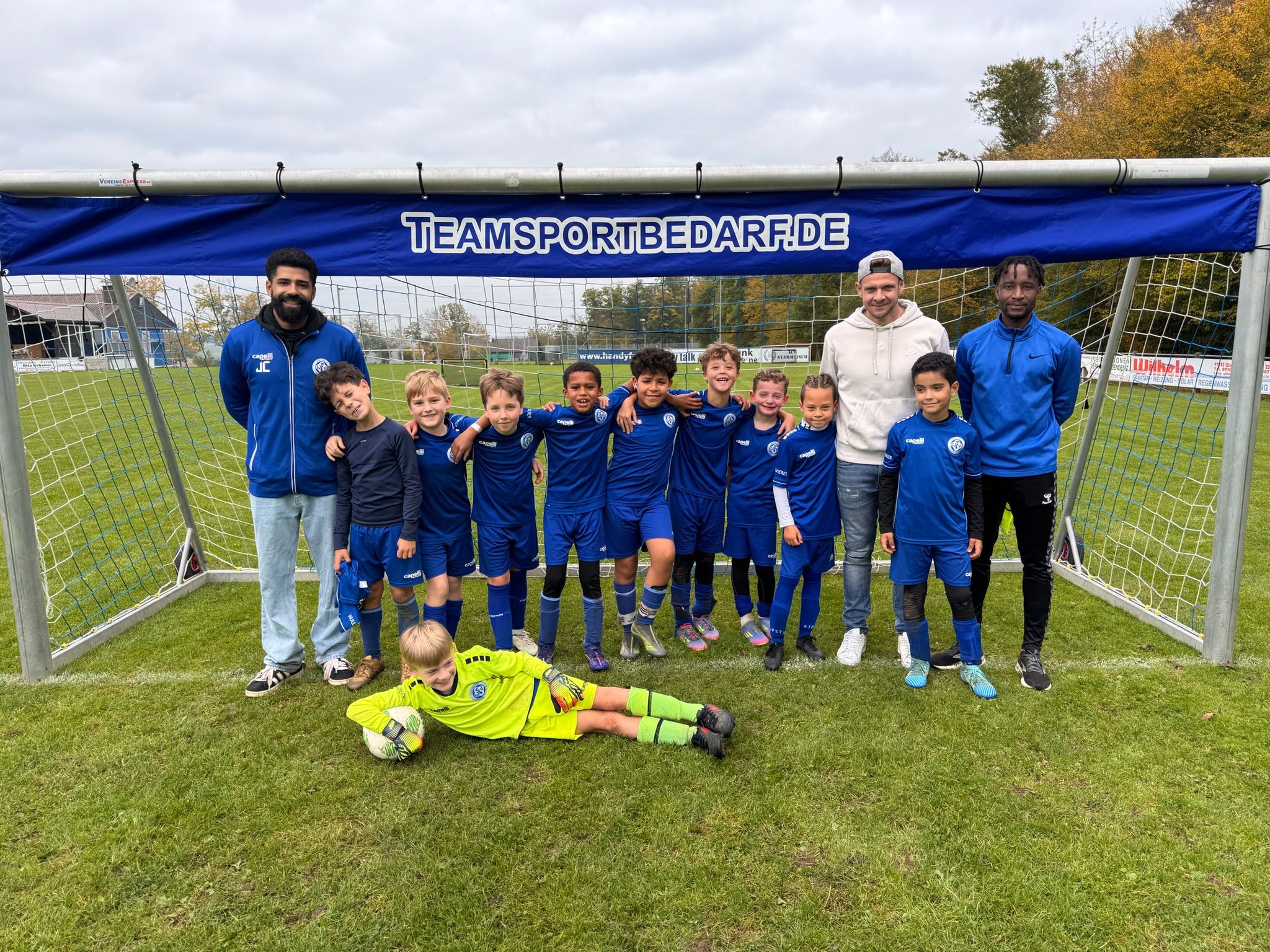 Kinderfußballmannschaft mit Trainern vor einem Tor mit Teamsportbedarf.de Banner auf einem Fußballplatz