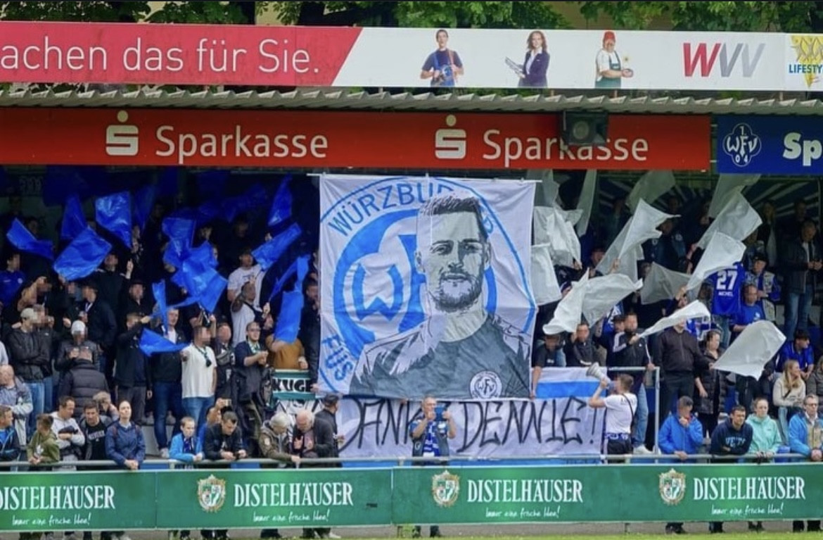 Fans des Würzburger FV mit blau-weißen Fahnen und Banner im Stadion vor Sparkasse-Werbung